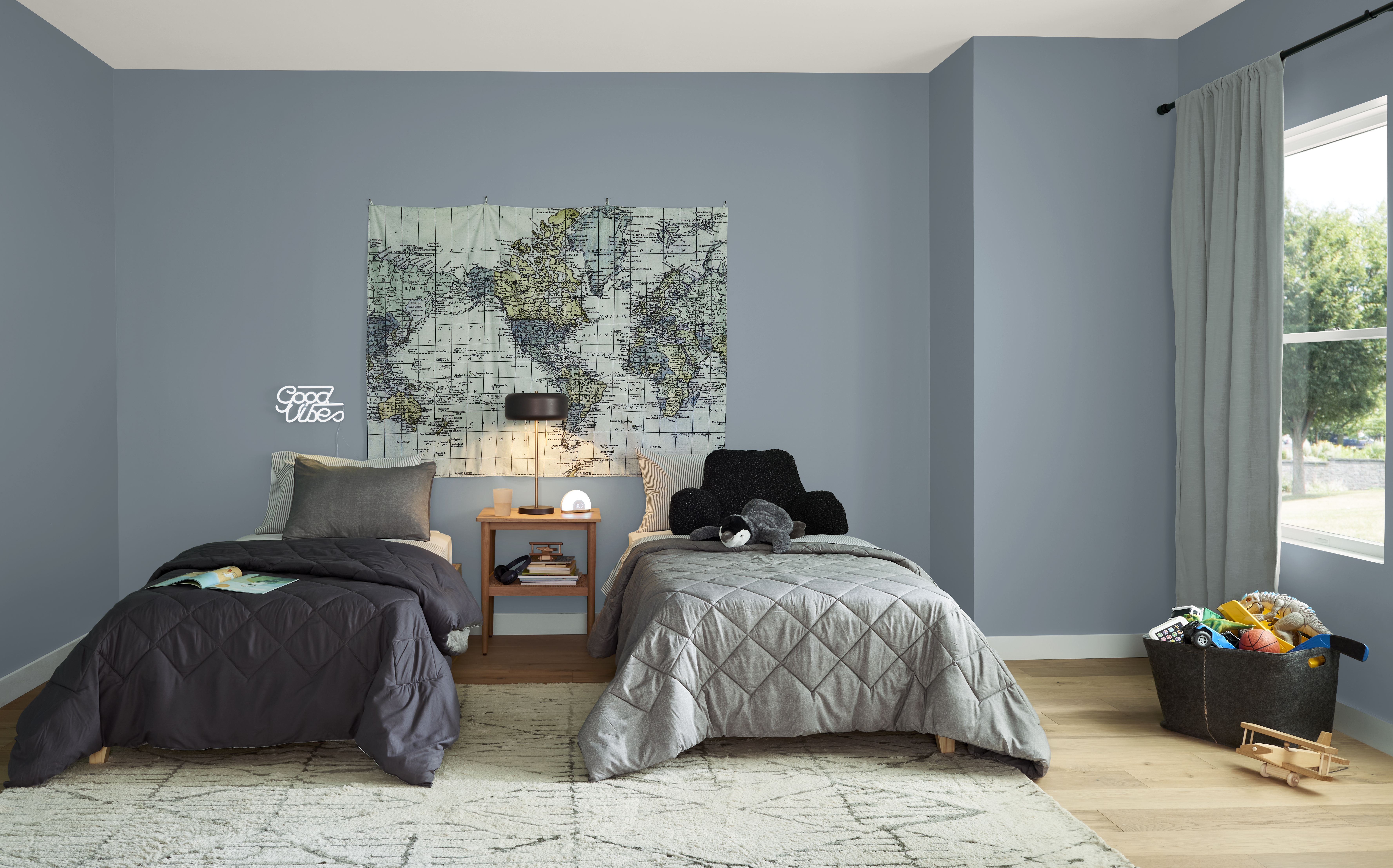 – A calm, neutral-toned kids’ bedroom with blue-grey walls, featuring two twin beds with textured grey bedding, a shared nightstand with a lamp, a world map wall art above the beds, and soft natural light from a window.