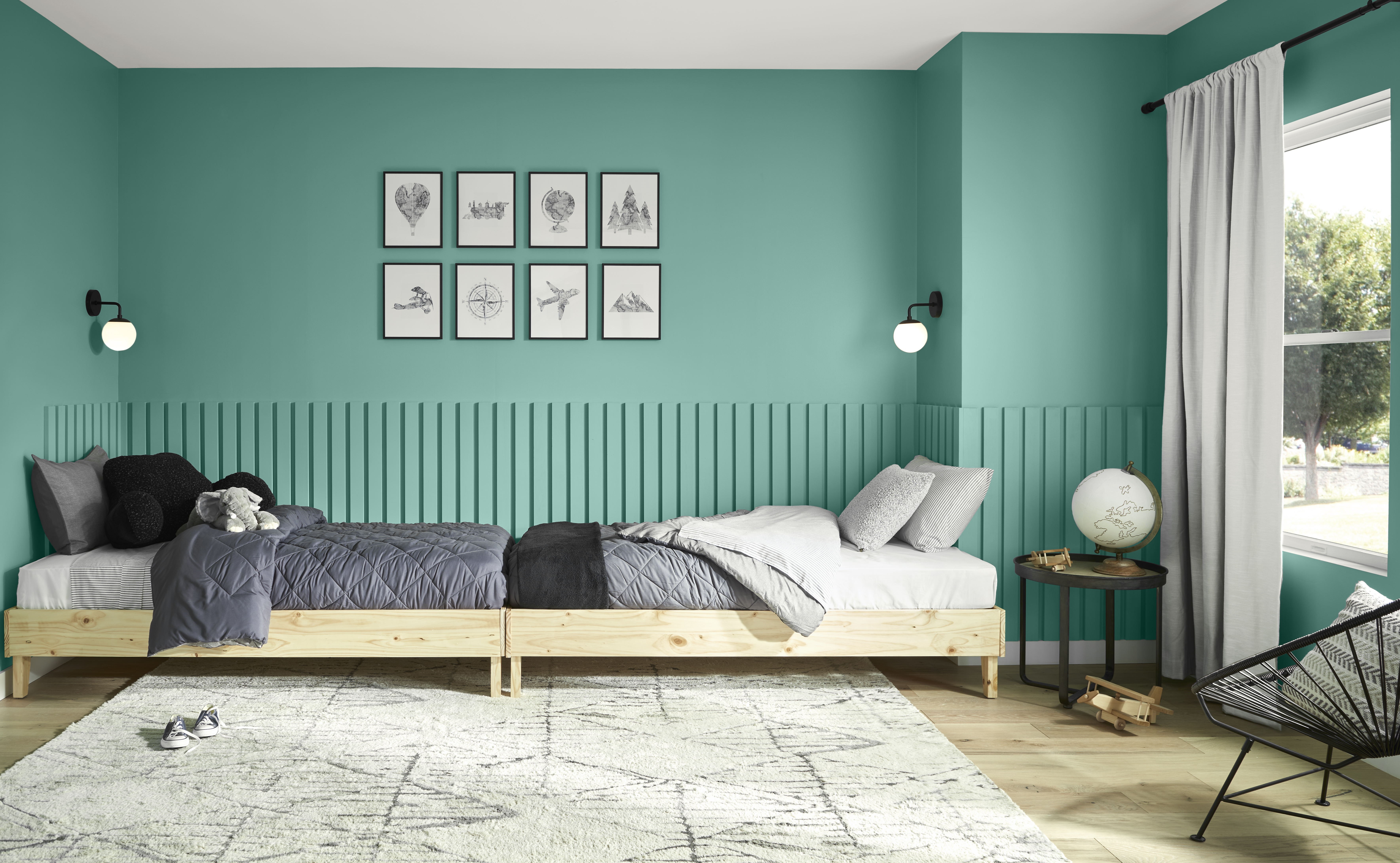 A modern kids’ bedroom with midtone green painted walls and paneled detail, featuring two low wooden twin beds with grey bedding, minimal black-and-white artwork above the beds, and natural light from a window creating a calm, cohesive space.