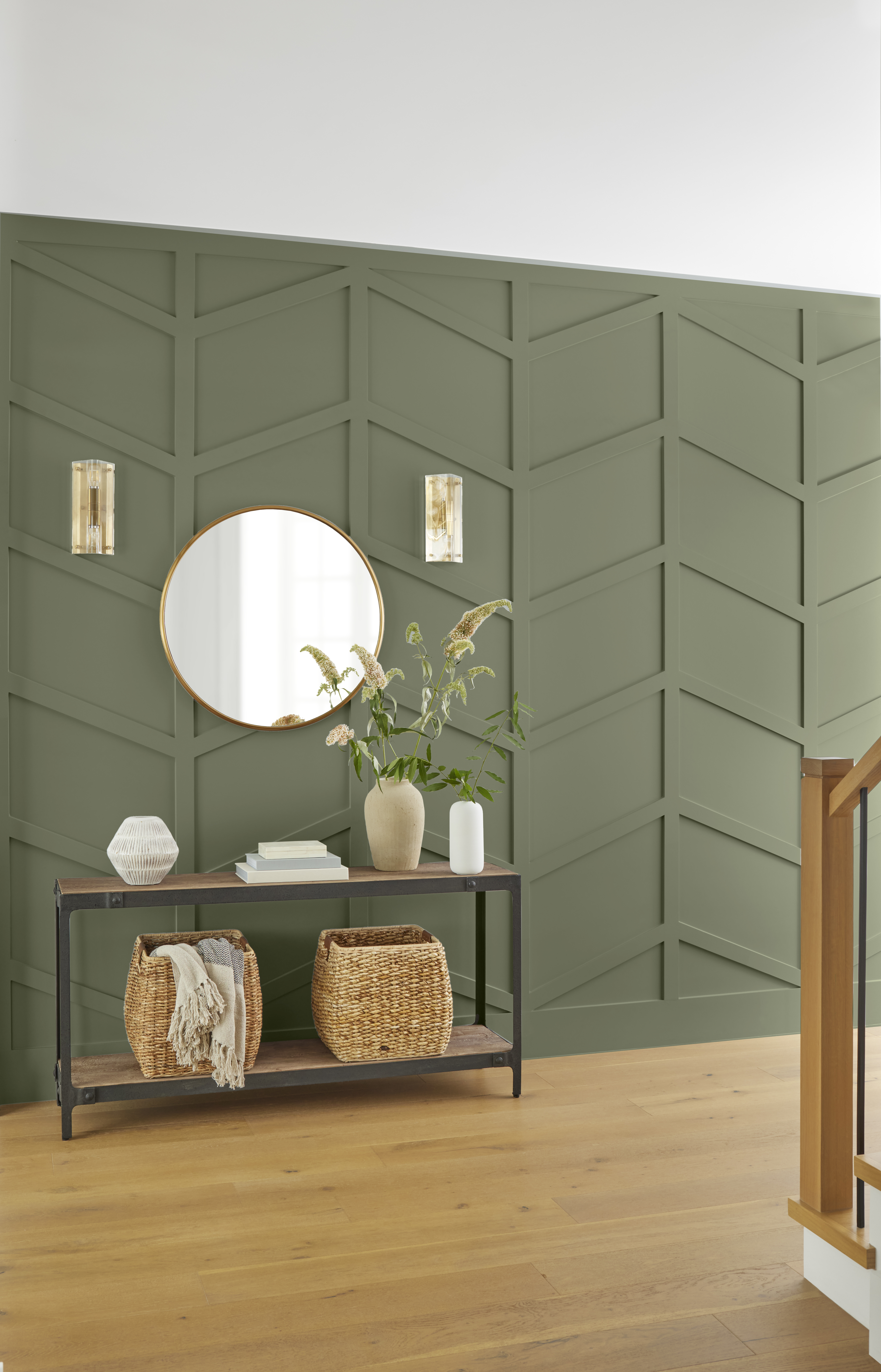 An entryway with geometric paneled walls painted in “Ecological,” featuring a round mirror, wall sconces, and a console table with woven baskets and décor.