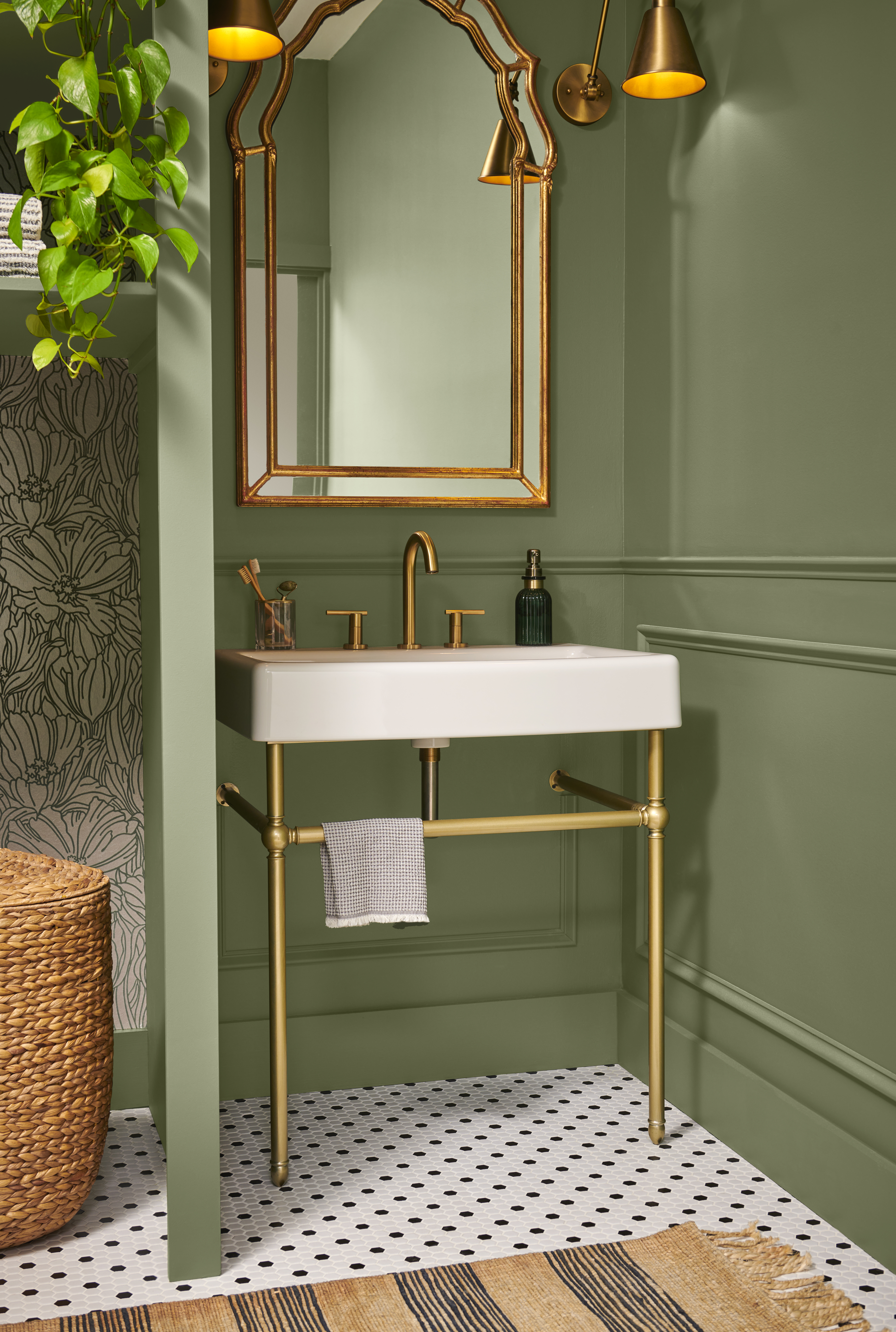 A small powder room with walls painted in “Ecological,” featuring a white pedestal sink with brass fixtures, a gold-framed mirror, and warm, modern accents.