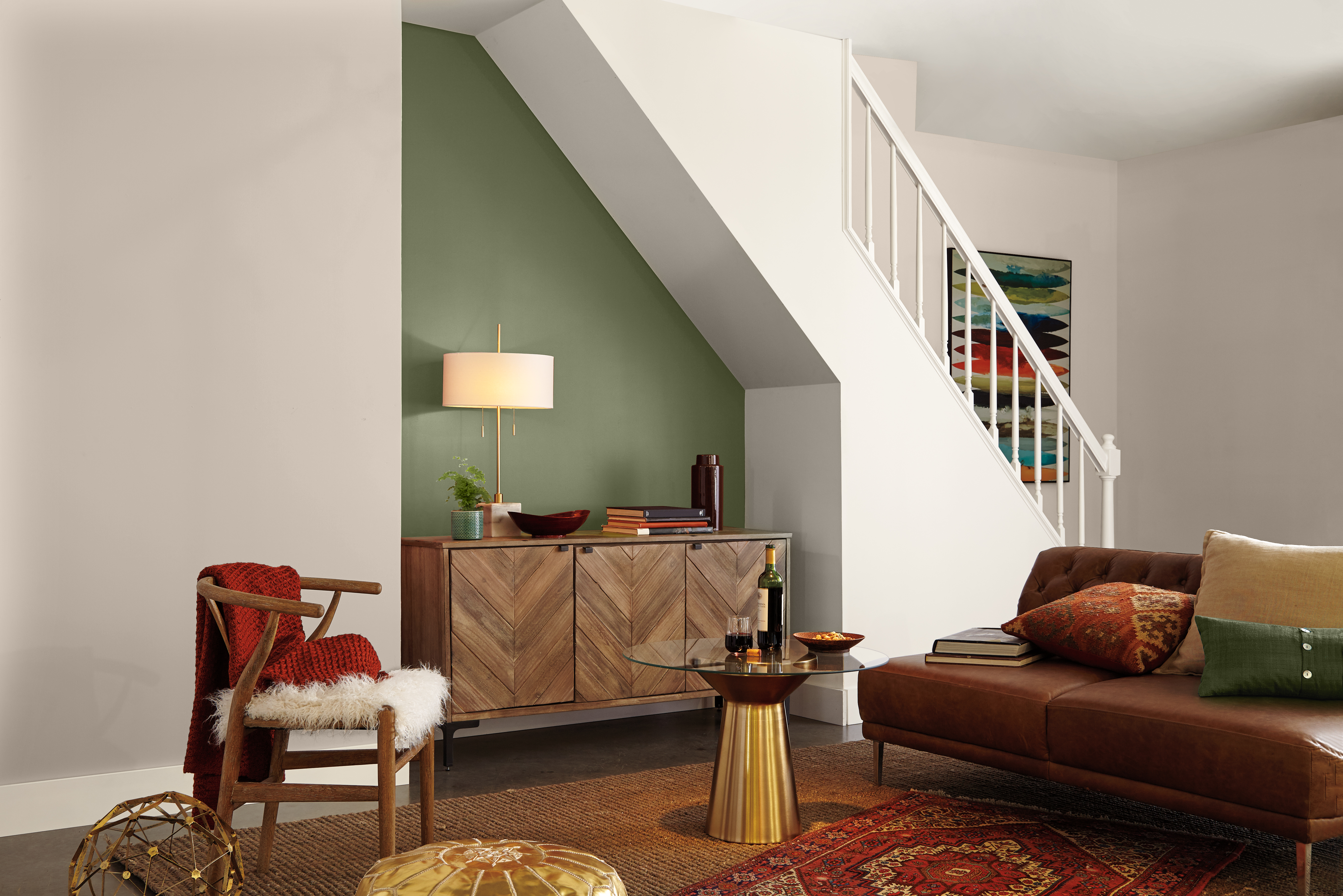 A basement living area painted with an earthy green paint called  “Ecological” as the accent wall under the staircase, styled with a wood sideboard, leather sofa, and warm, layered décor.