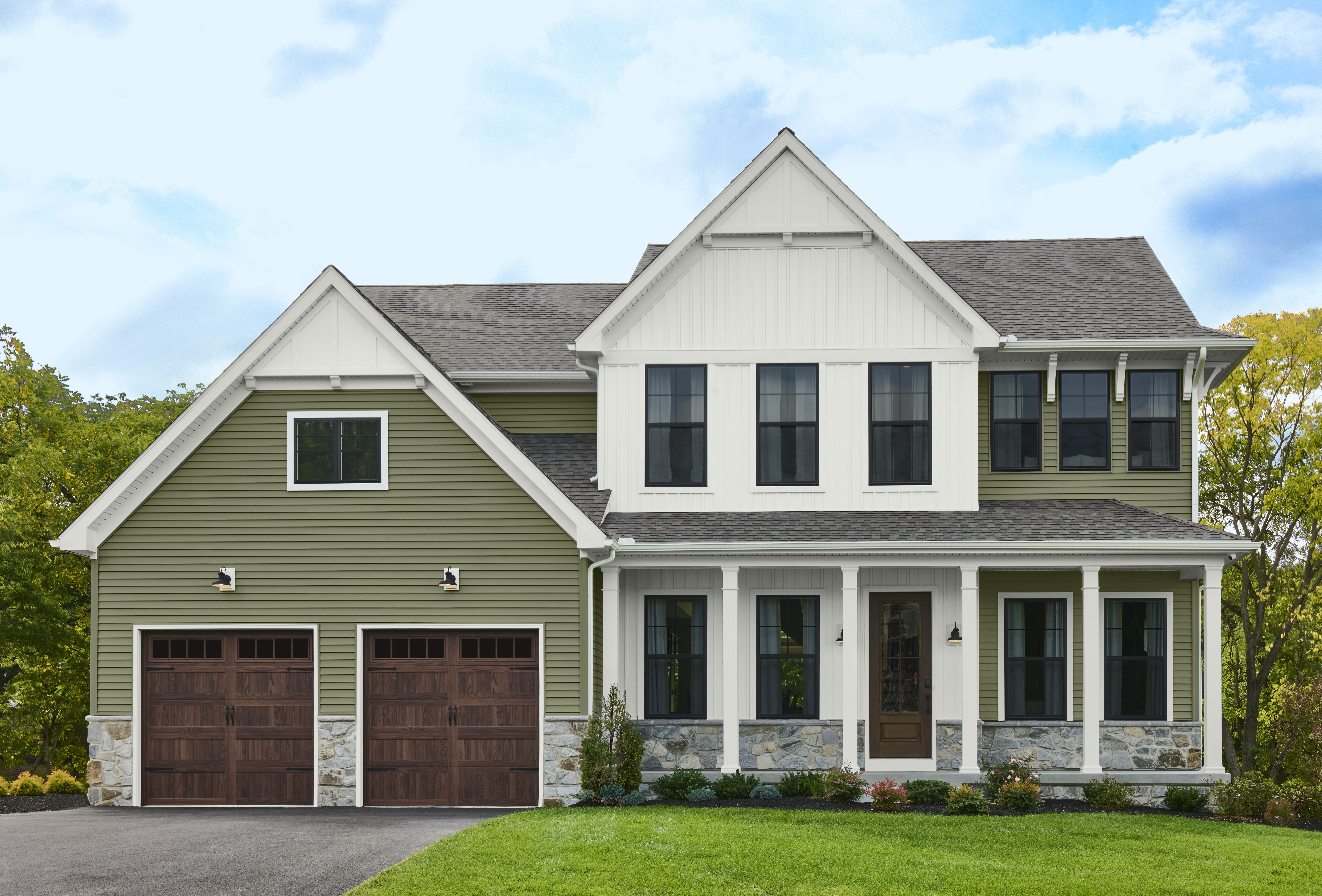 A two-story green and white farmhouse-style home with a double garage, front porch, stone accents, and green lawn.