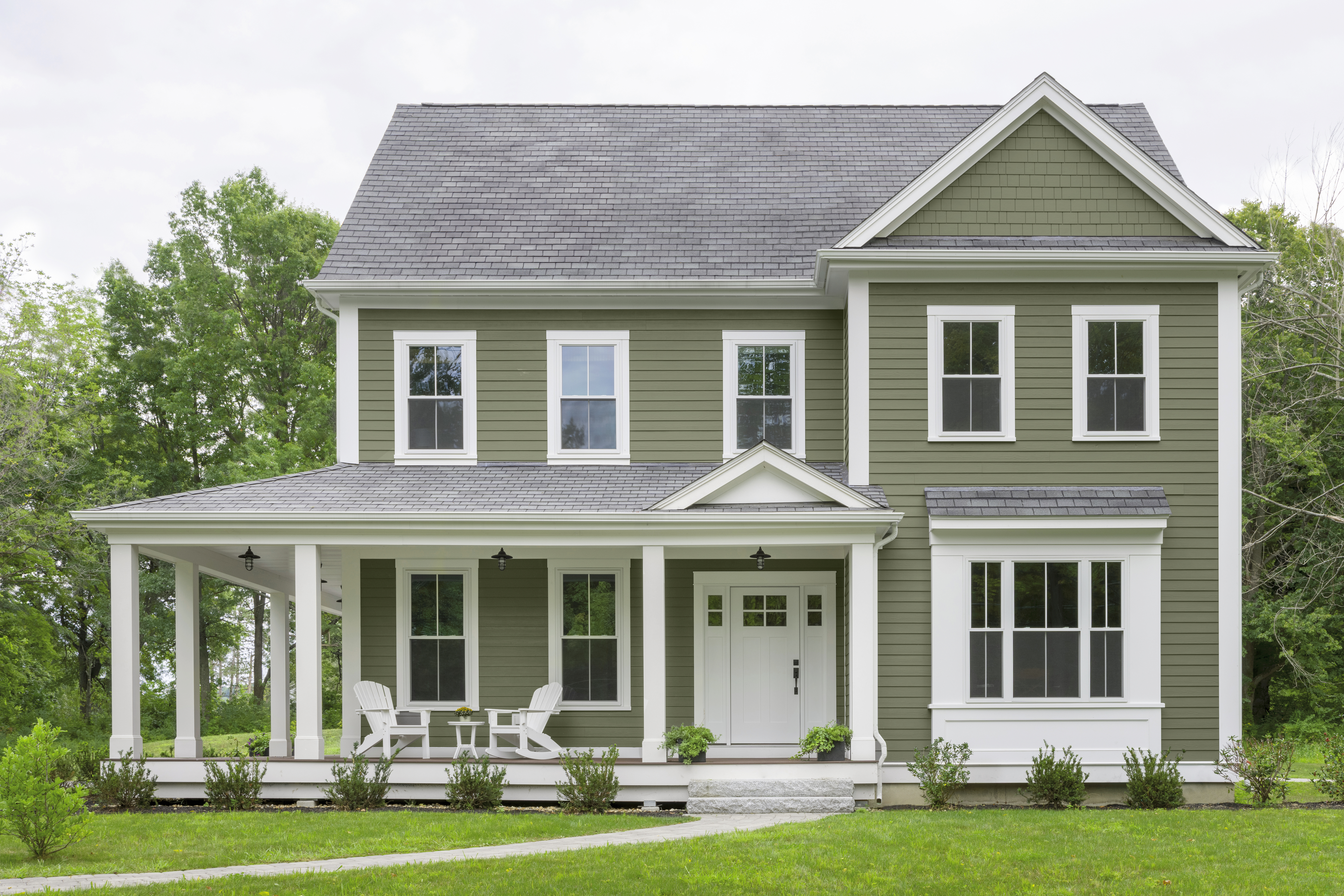 A two-story green coloured farmhouse-style home with white trim, a wraparound porch, and a green front lawn.