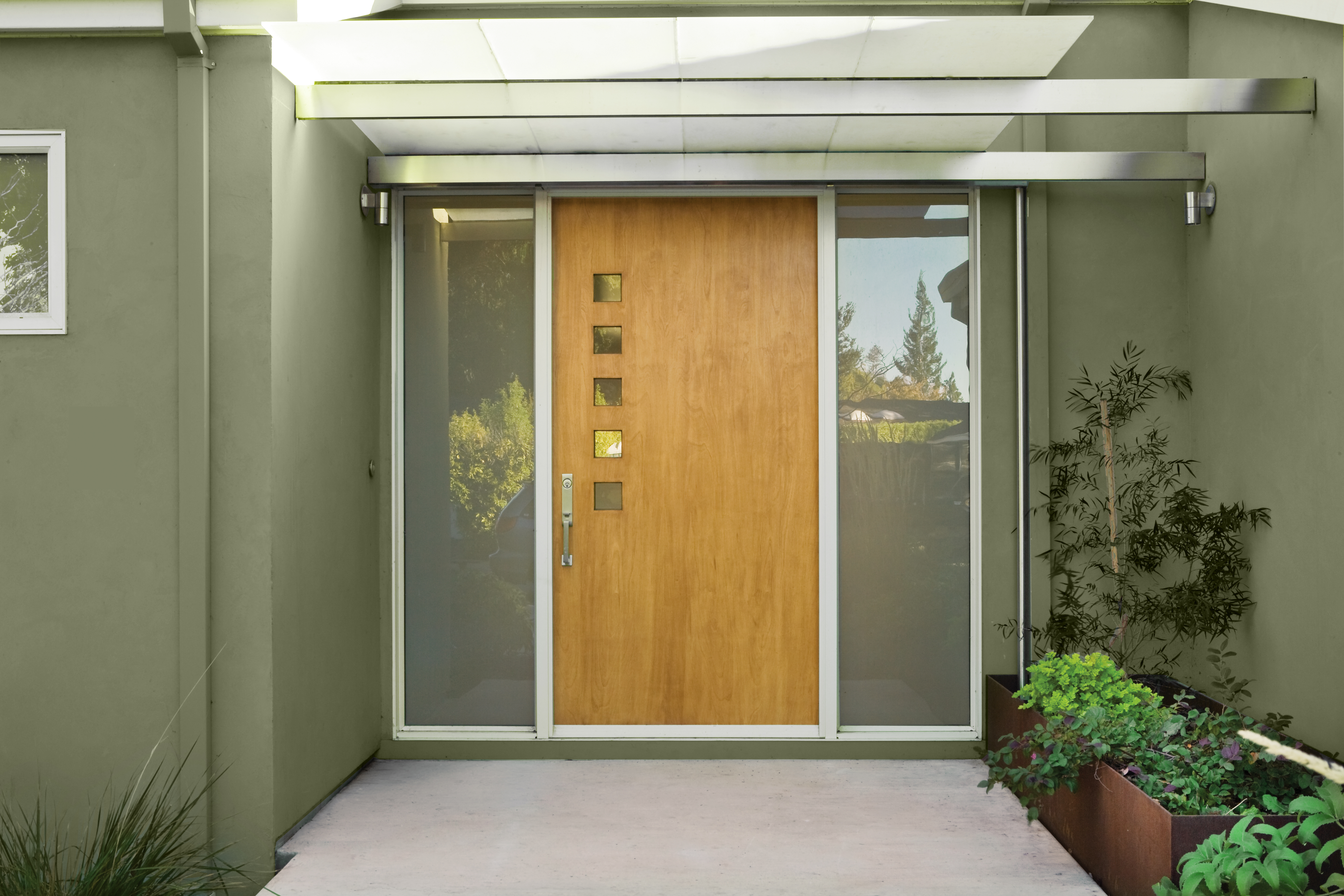 A Modern entryway with a light wood front door, frosted glass sidelights, metal awning, and potted plants. The house body is covered in green stucco.