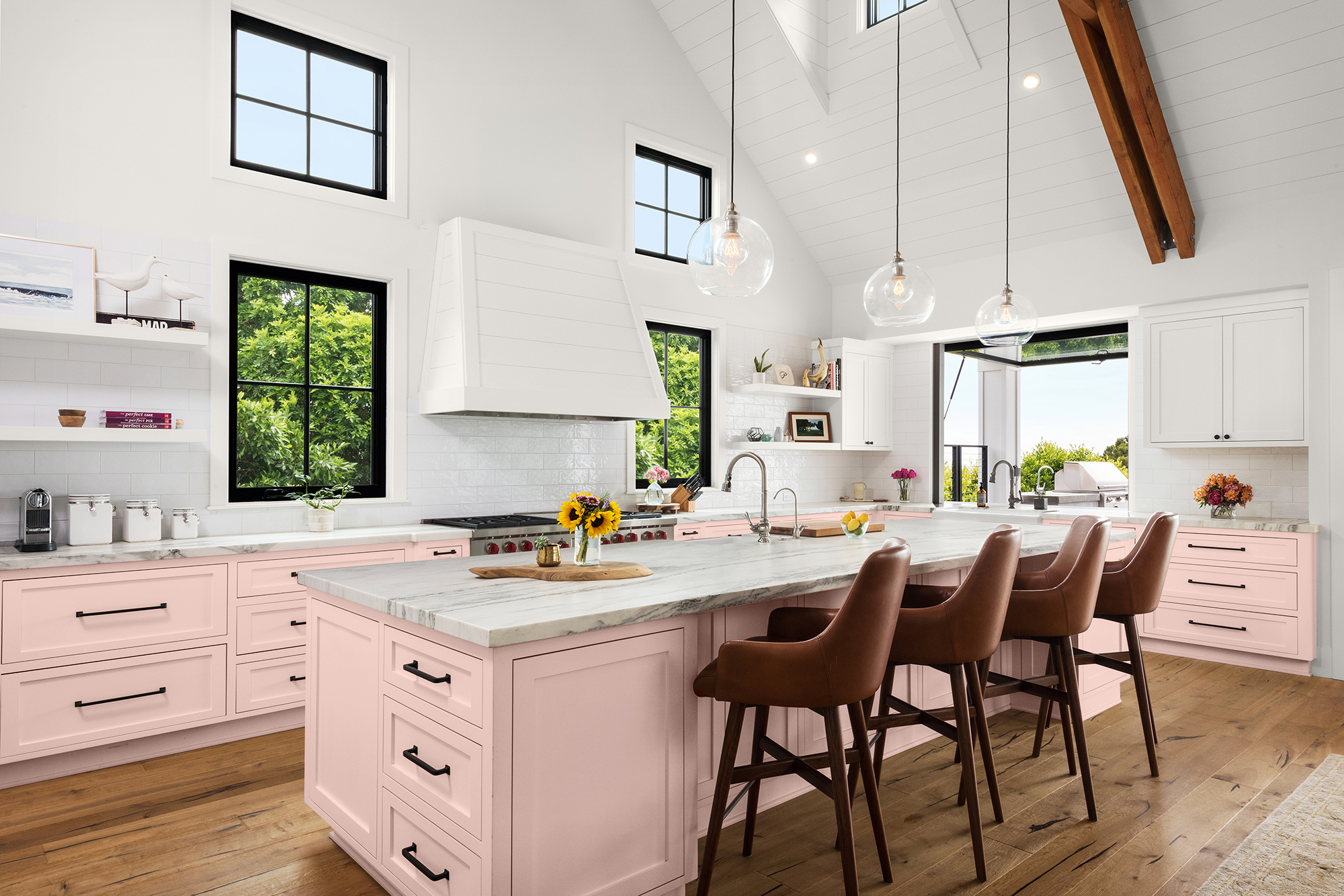 A pink kitchen with pastel cabinets, marble countertops, black-framed windows, pendant lights, and a large island with brown bar stools.