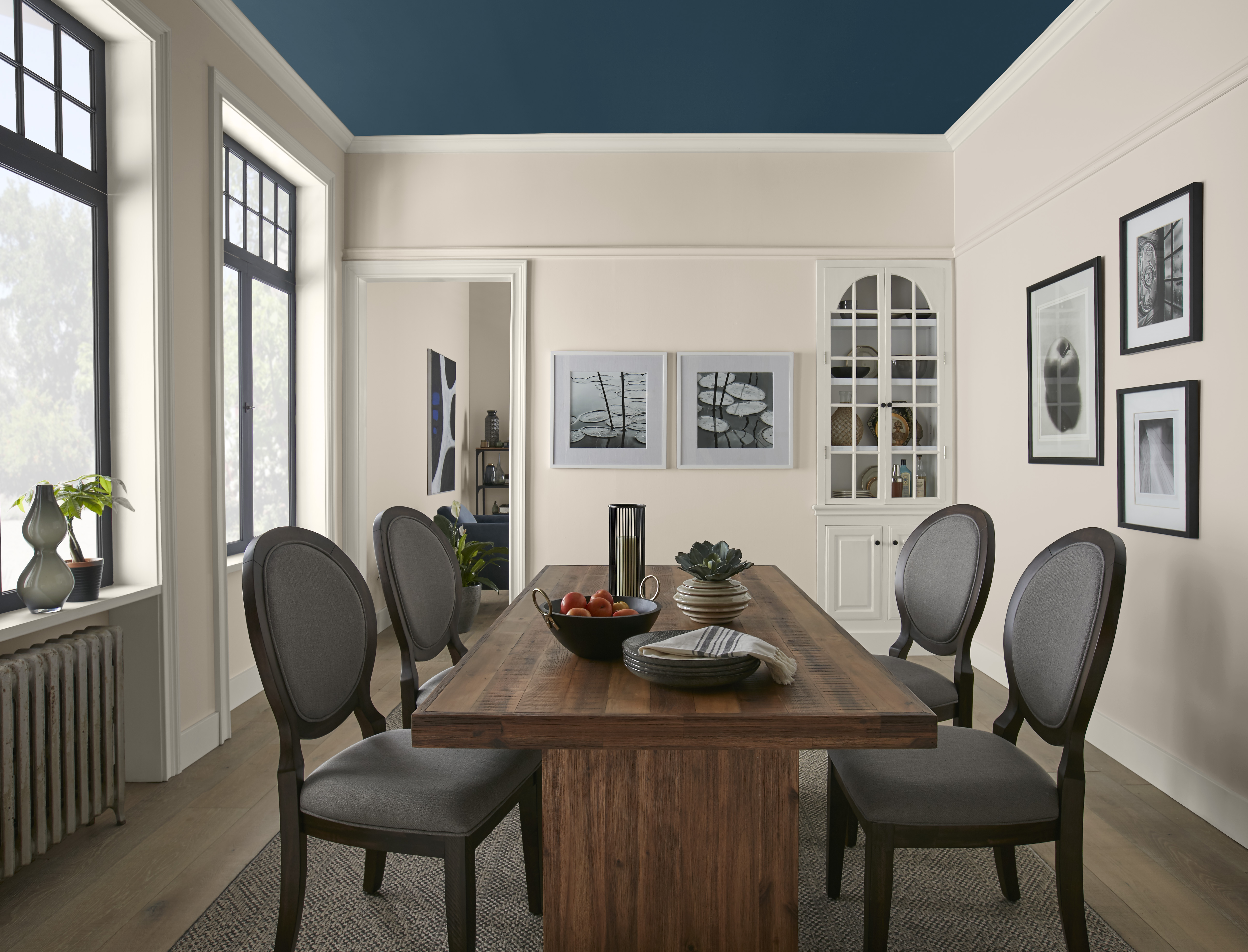 A dining room with Wheat Bread walls, crisp white trim and a dark blue ceiling, featuring a wood dining table, grey upholstered chairs, tall black-framed windows and framed wall art.