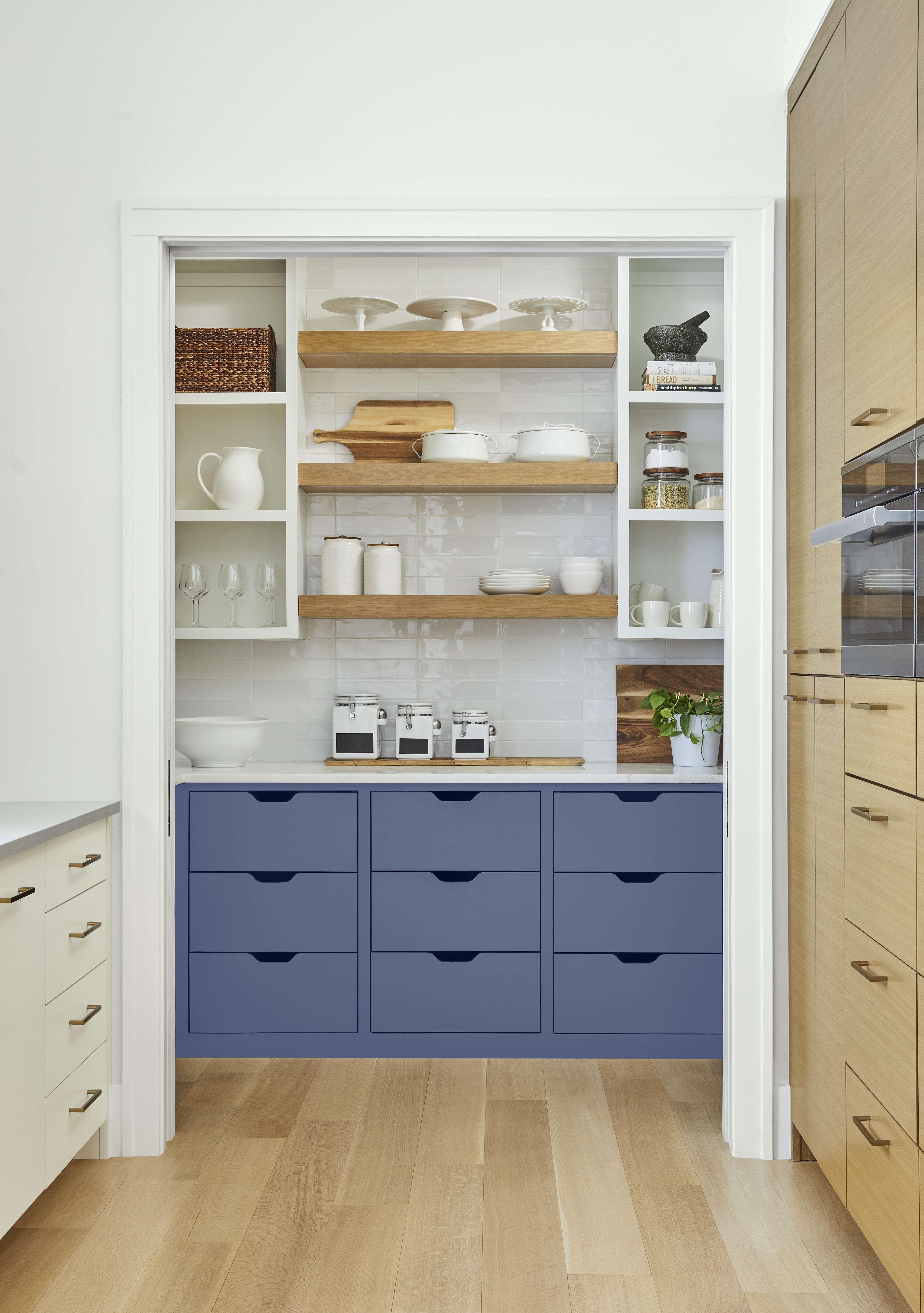 A pantry nook with blue-purple lower cabinets that create a surprise pop of colour, paired with open wood shelving, white built-in storage, natural wood cabinetry, and light wood floors.