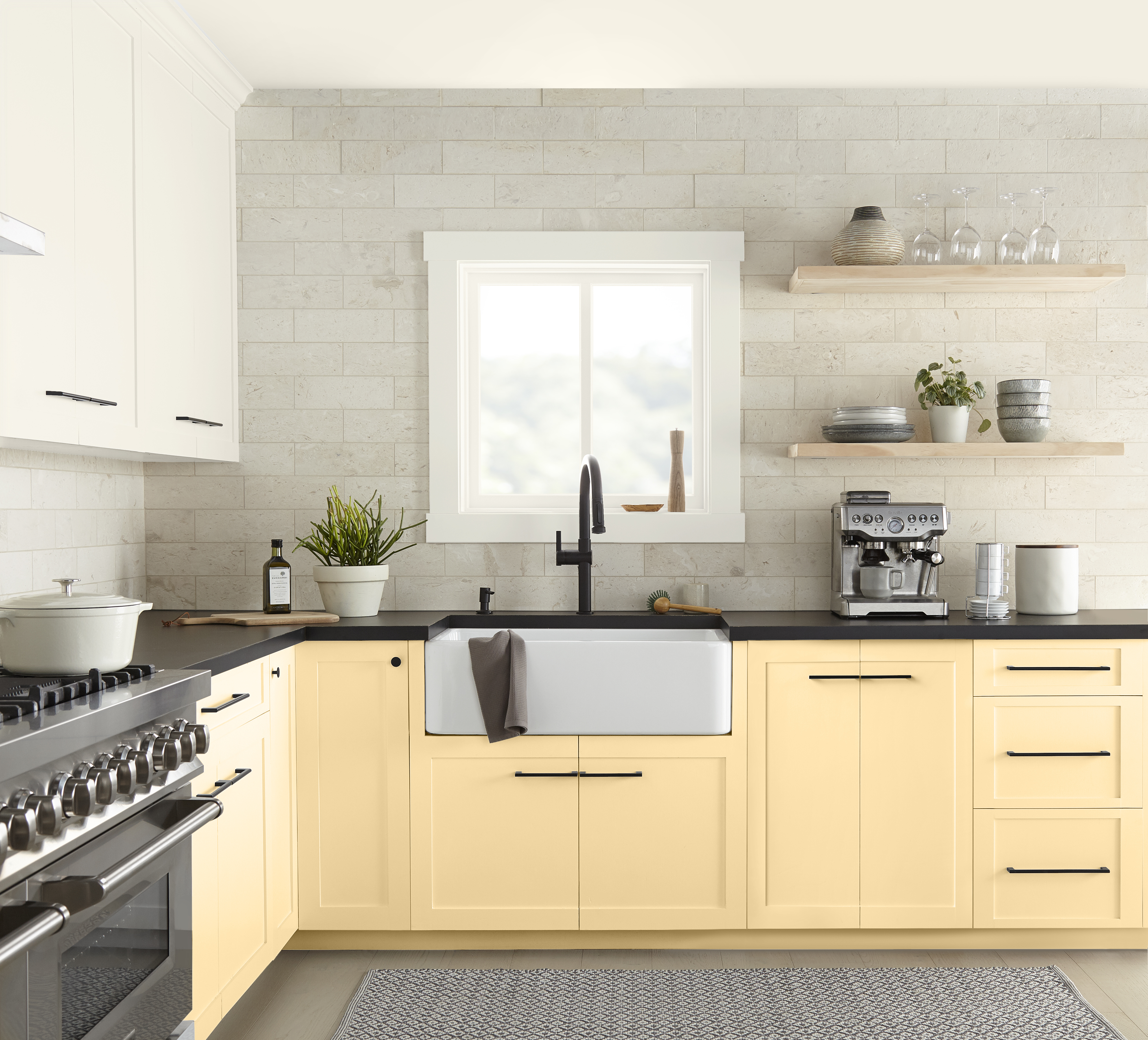 A bright kitchen with soft yellow paint colour on the lower cabinets, a farmhouse sink, black hardware, and a light stone backsplash. The soft yellow cabinetry helps the small kitchen feel larger, brighter, and more inviting by reflecting light and creating a warm, airy atmosphere.