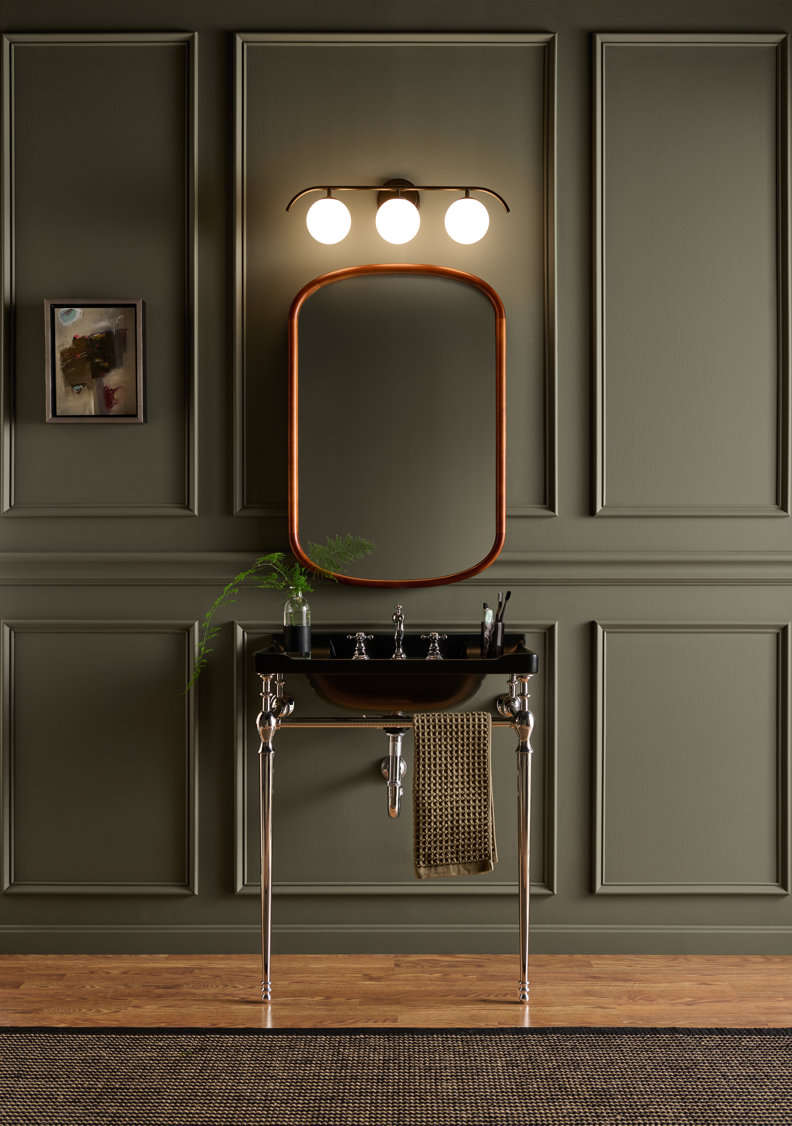 A sophisticated small bathroom with decorative moulding painted in BEHR Grounded, featuring a black console sink, rounded mirror, globe vanity lights, and warm wood flooring.