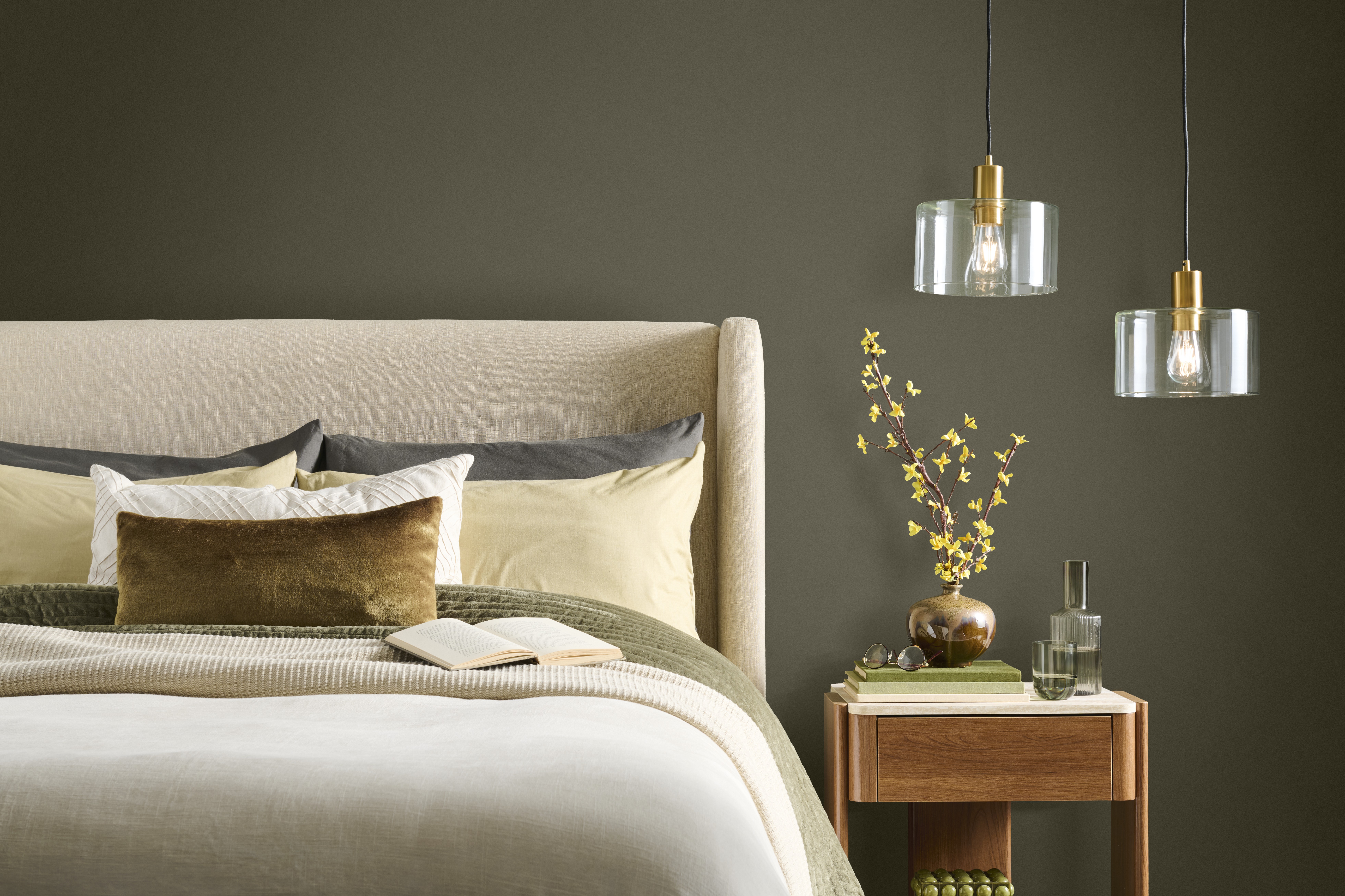A contemporary and serene bedroom with an accent wall painted in BEHR Grounded, featuring a neutral upholstered bed, layered bedding, a wooden nightstand, and glass pendant lights.