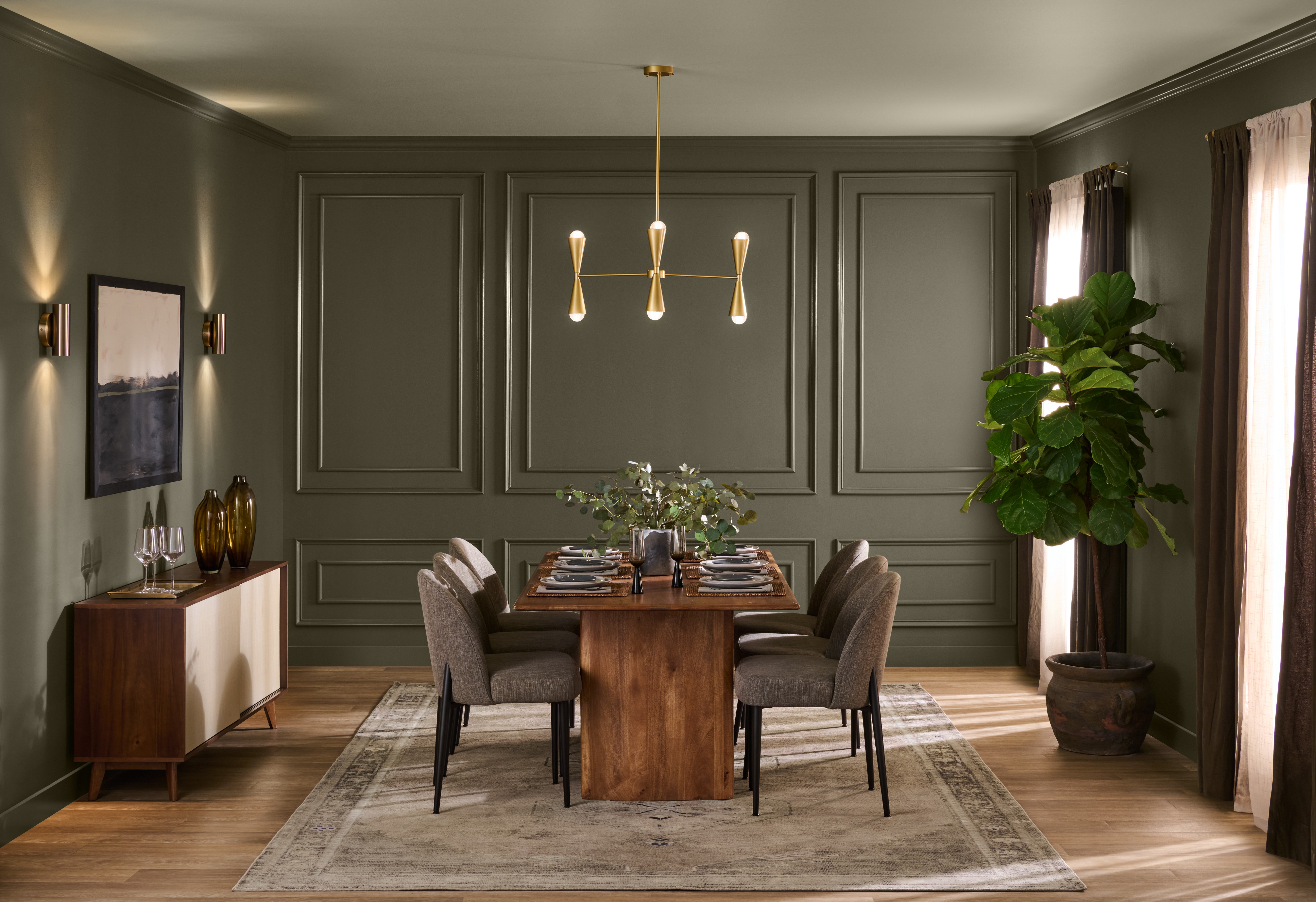 A sophisticated dining room with walls painted in BEHR Grounded, featuring decorative wall moulding, a wooden dining table, upholstered chairs, a modern brass chandelier, and warm natural light.