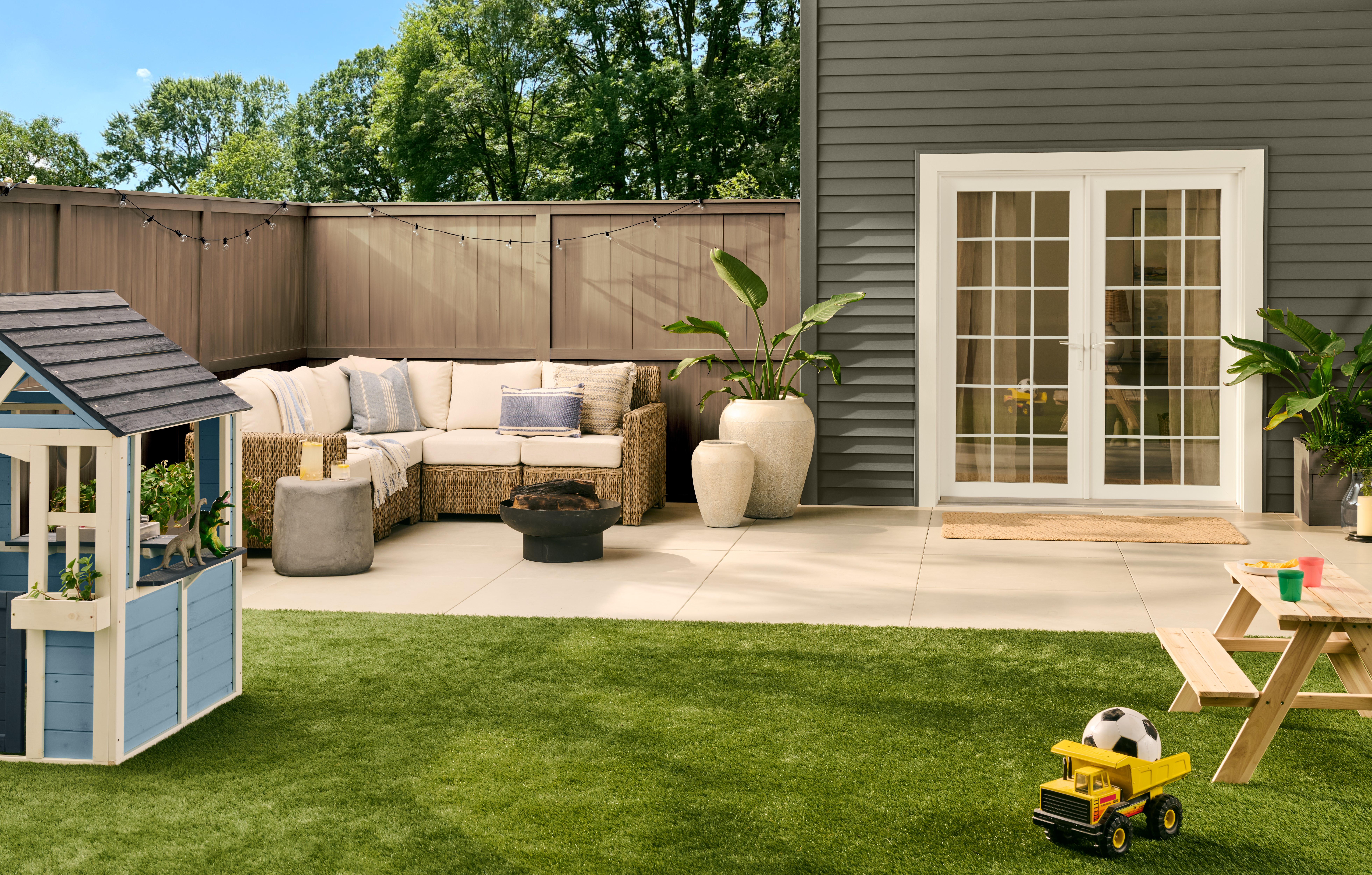 A family-friendly backyard with exterior siding painted in BEHR Grounded, featuring a paved patio, outdoor seating, French doors, a green lawn, and a children’s play area.
