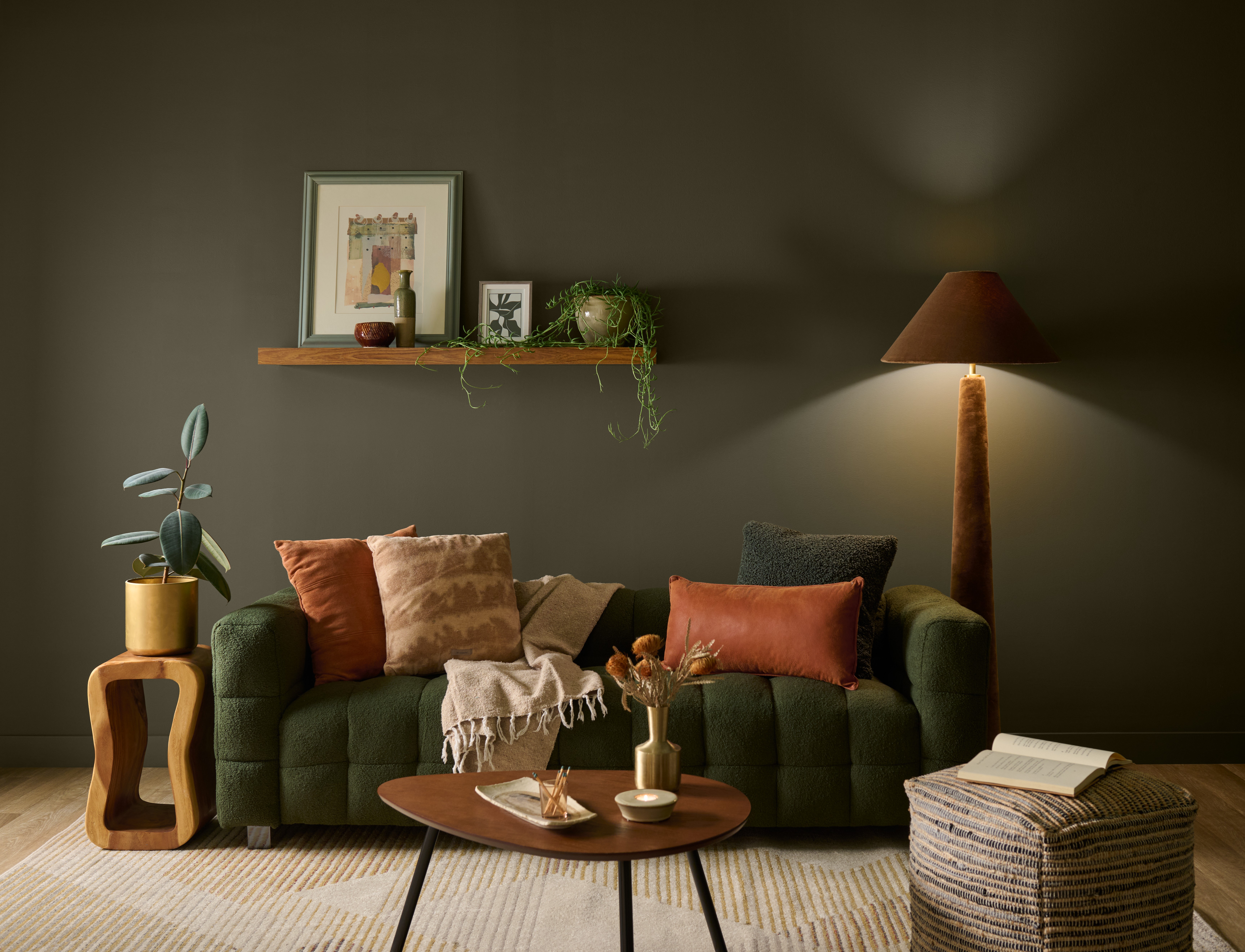 A cozy living room with walls painted in BEHR Grounded, featuring a deep green sofa, warm rust-coloured pillows, a wooden coffee table, layered natural textures, and soft ambient lighting.