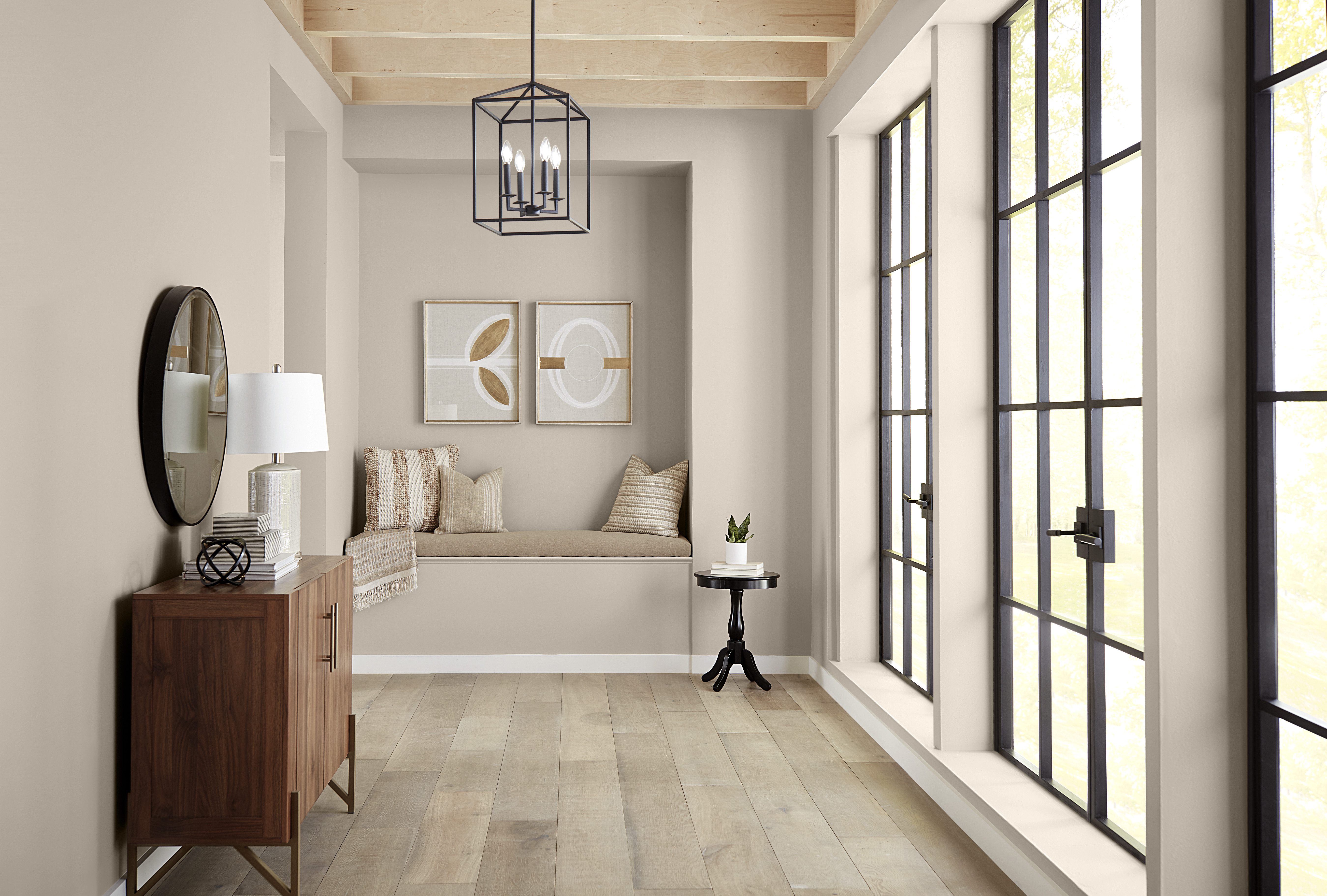 A light-filled hallway with walls painted in Wheat Bread, creating a warm neutral backdrop for black-framed windows, pale wood flooring, exposed ceiling beams and a cozy built-in seating nook.