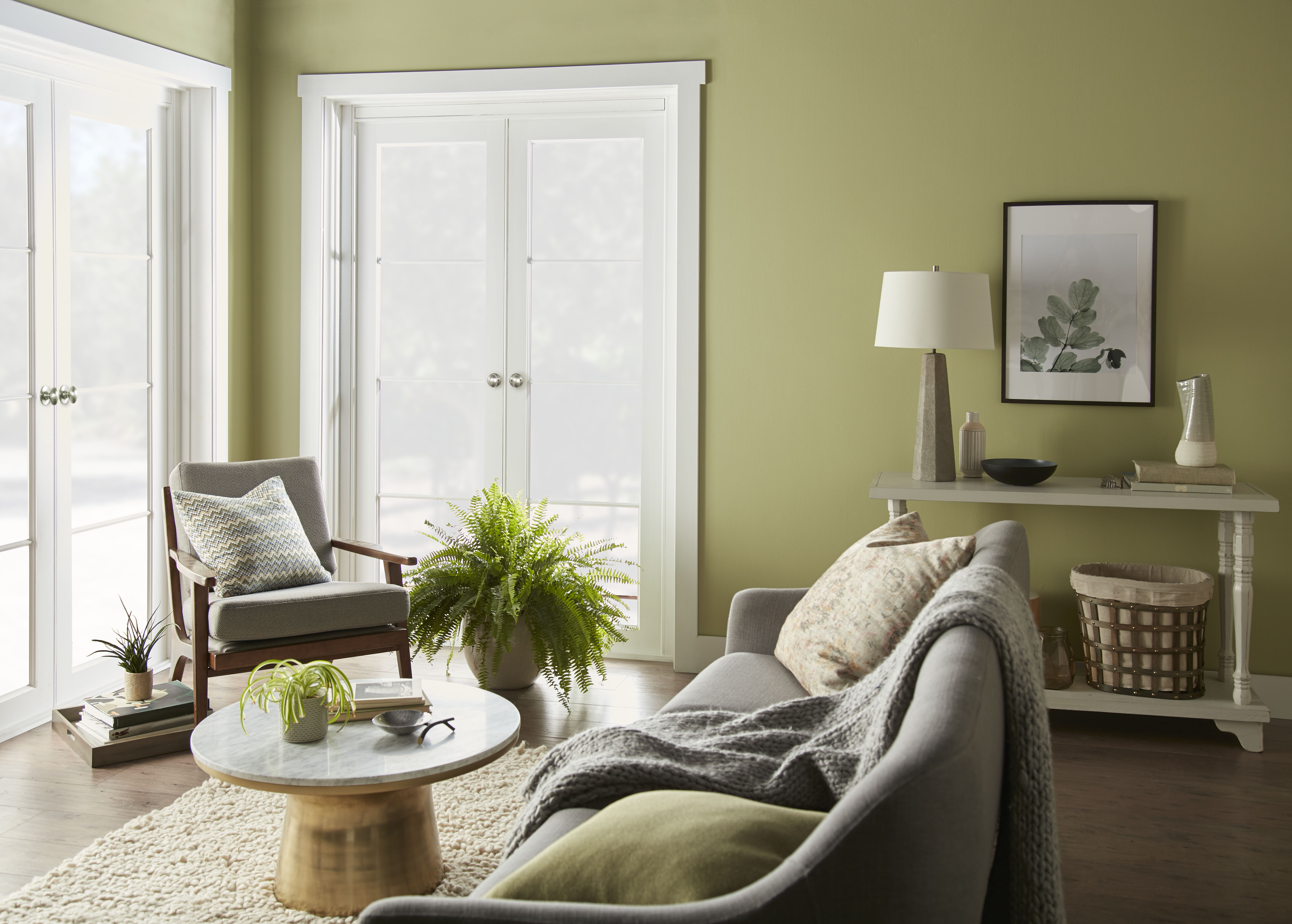 Living room with Fresh Artichoke green wall paint, natural wood furniture, soft neutral upholstery, indoor plants, and layered textures for a restorative earthy colour palette.