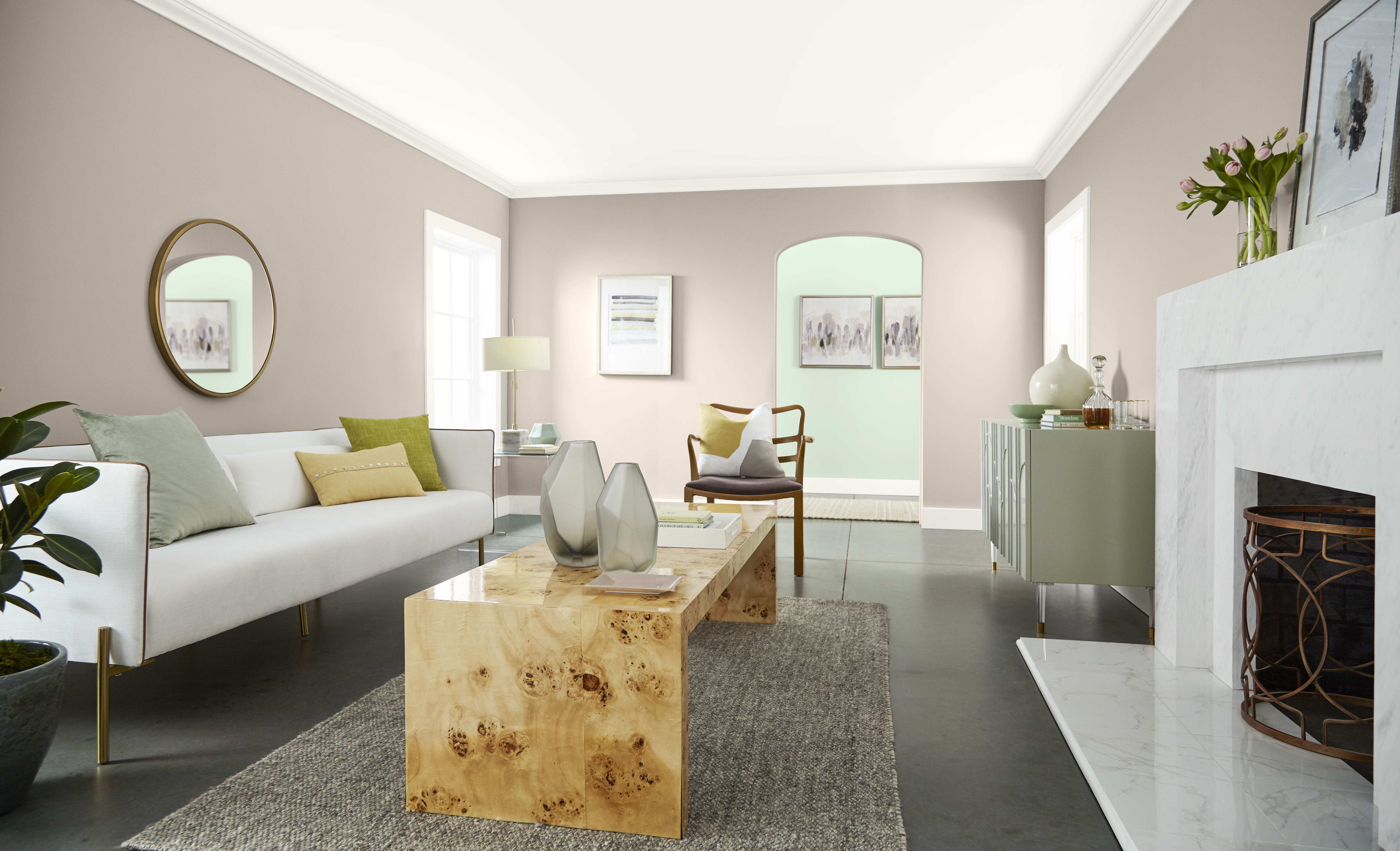 A living room with walls painted in Wheat Bread, featuring a white sofa, natural wood coffee table, grey area rug, fireplace and soft green décor