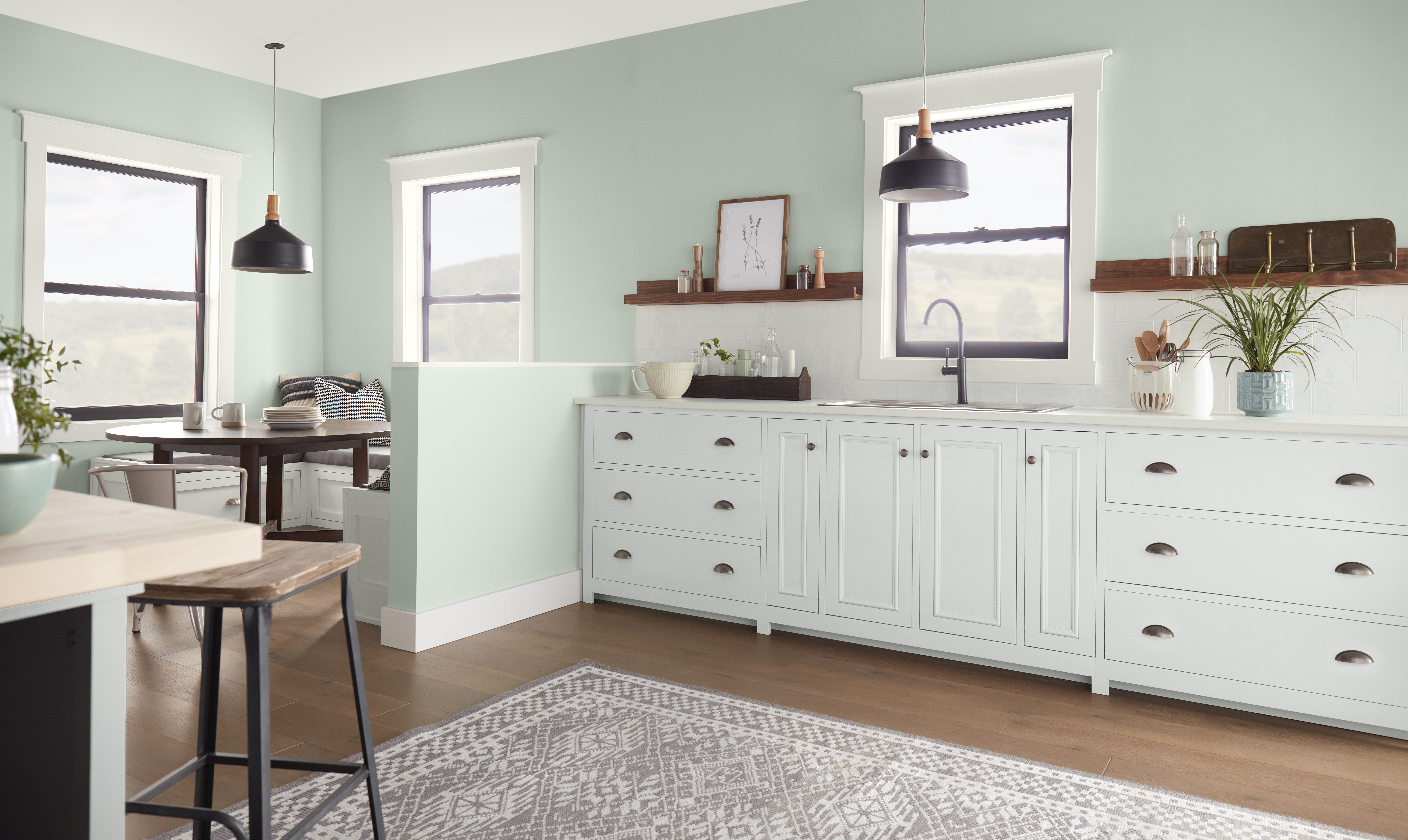 A bright kitchen with soft green walls, white cabinetry, black pendant lights, wood shelving, and large windows. The green colour gives the kitchen a fresh, calming feel while adding subtle contrast to the white cabinets and natural wood tones.