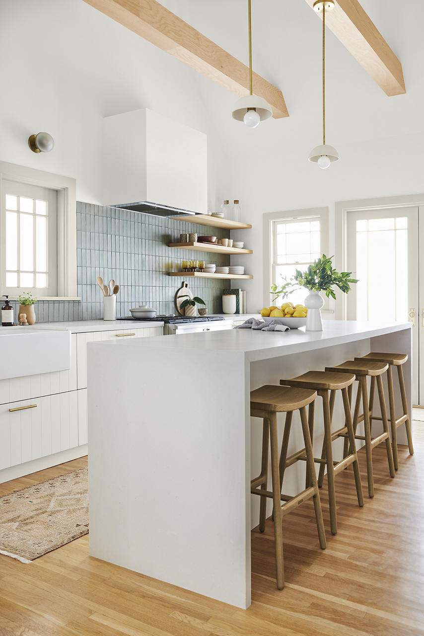 A layered, textured modern organic farmhouse kitchen with white walls and white cabinetry, a large light grey island, light wood beams, open shelving, wooden bar stools, pale blue tile on the backsplash, and warm natural wood floors.