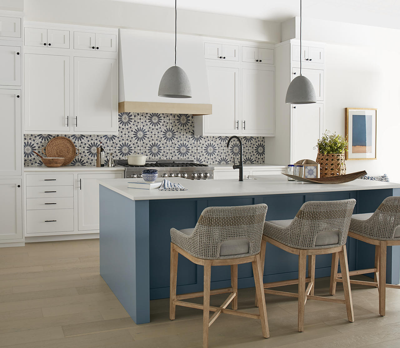 A white kitchen with a dusty and top-selling blue paint colour on the island, blue patterned tile backsplash, white cabinets, grey pendant lights, woven bar stools, and light wood floors.