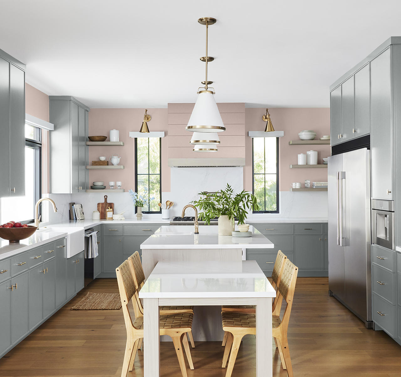 A bright kitchen with grey-blue cabinets, brass hardware, white countertops, a soft pink accent wall, wood dining chairs, and warm natural light. The grey-blue cabinetry brings a calm, sophisticated feel to the space, while the soft pink wall adds warmth and softness, creating a balanced kitchen that feels both fresh and inviting. Together, the colours add depth and character to this kitchen design.