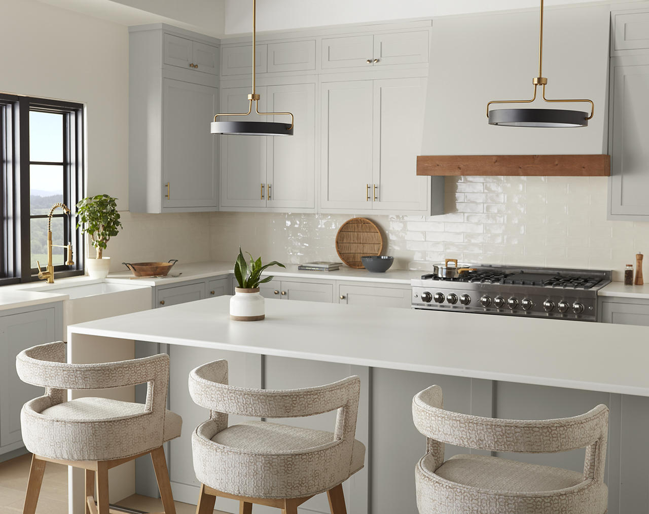 A white and light grey kitchen with warm natural wood range trim, brass hardware, brass pendant lights, white countertops, and textured neutral bar stools. The wood tones warm up the grey cabinetry, while brass hardware adds a modern, polished finish.