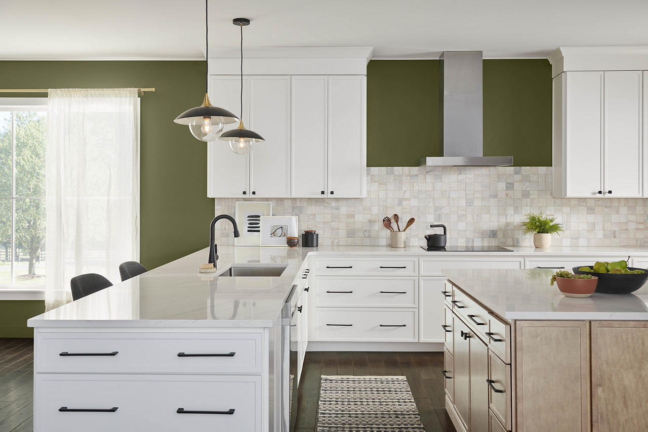A large and expansive kitchen with olive green walls, white cabinetry, black hardware, light wood cabinetry, and white quartz countertops. The olive-green colour adds natural warmth and earthy sophistication, creating a grounded, welcoming atmosphere. Paired with crisp white cabinets and light wood tones, it brings depth, contrast, and a timeless organic feel to the space.