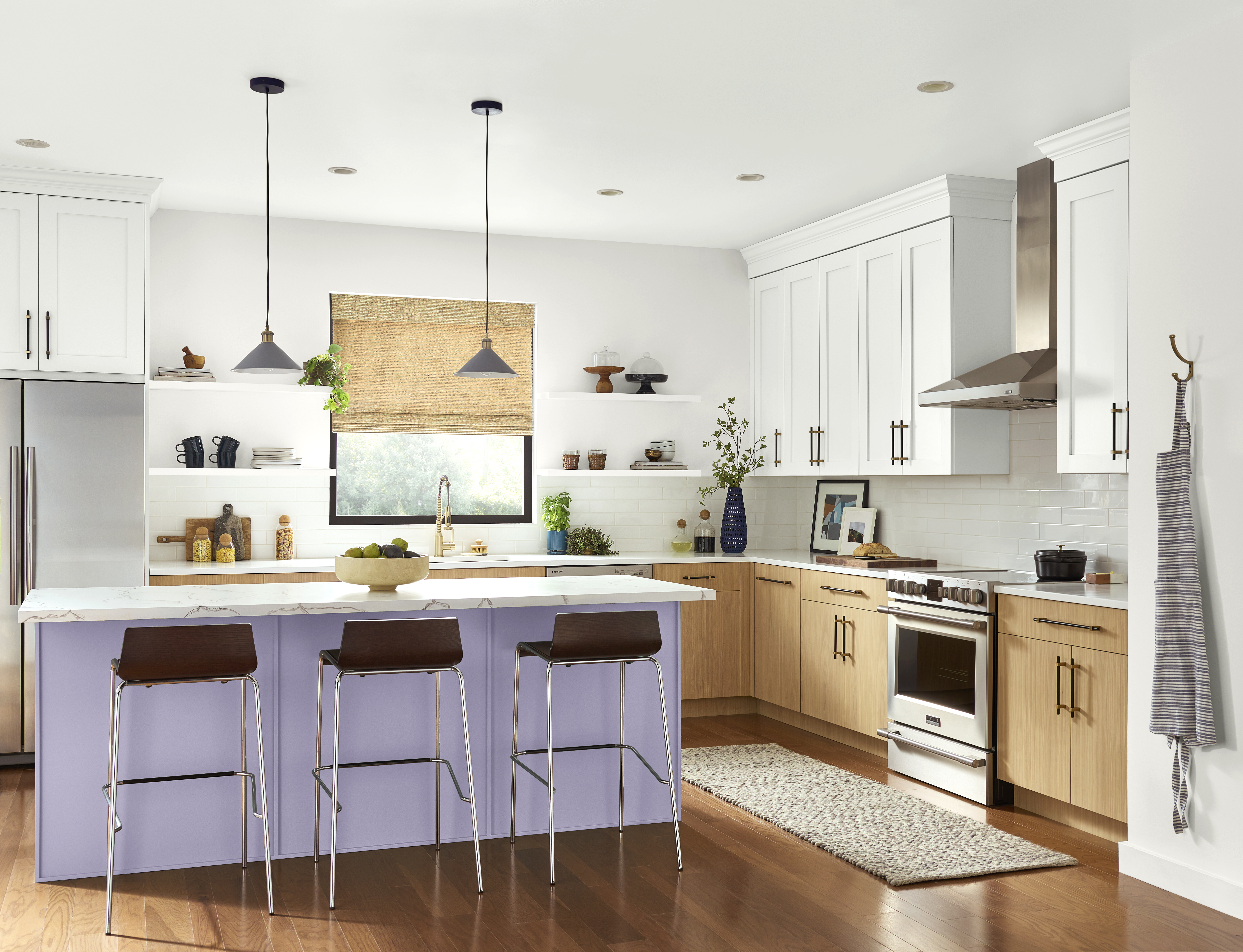 A kitchen with a lavender island, white upper cabinets, natural wood lower cabinets, white countertops, pendant lights, and warm wood floors. The soft purple shade makes the kitchen feel soft and approachable. Paired with crisp white countertops and natural wood cabinetry, this light purple feels fresh, balanced, and easy to use in everyday kitchen design.