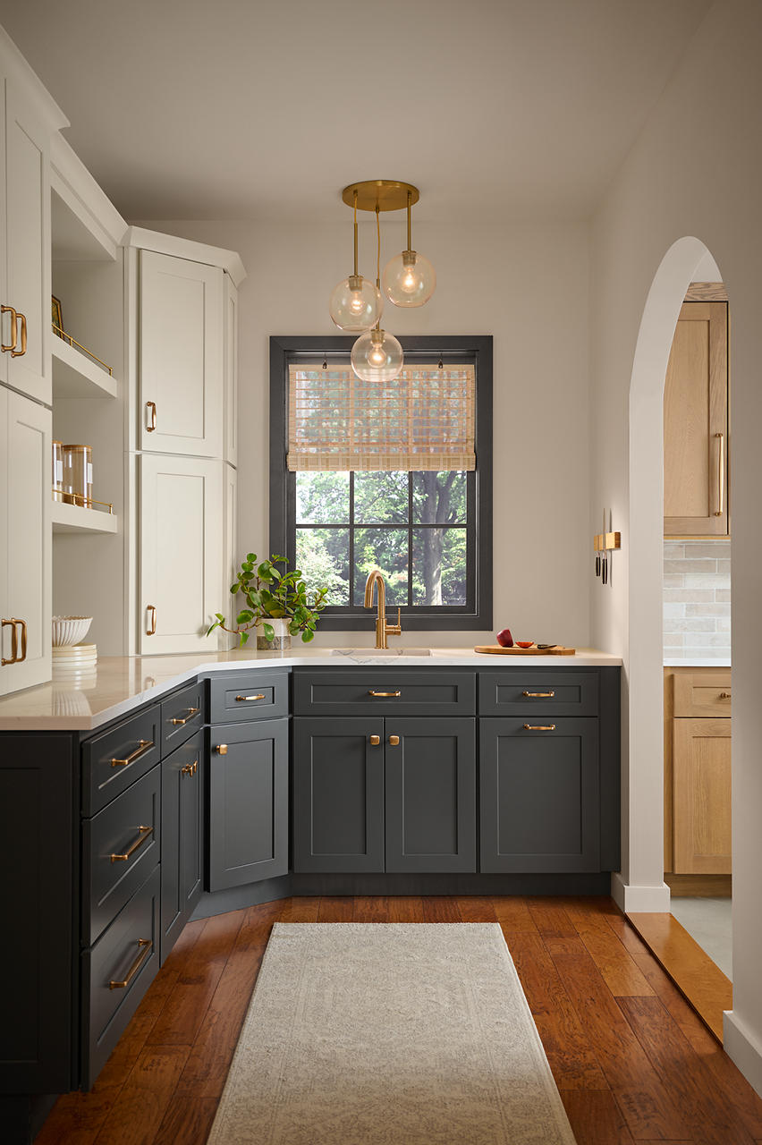 A walk-in pantry with walls painted in Wheat Bread, featuring dark lower cabinets, white built-in shelving, brass hardware, warm wood flooring and an arched doorway. 
