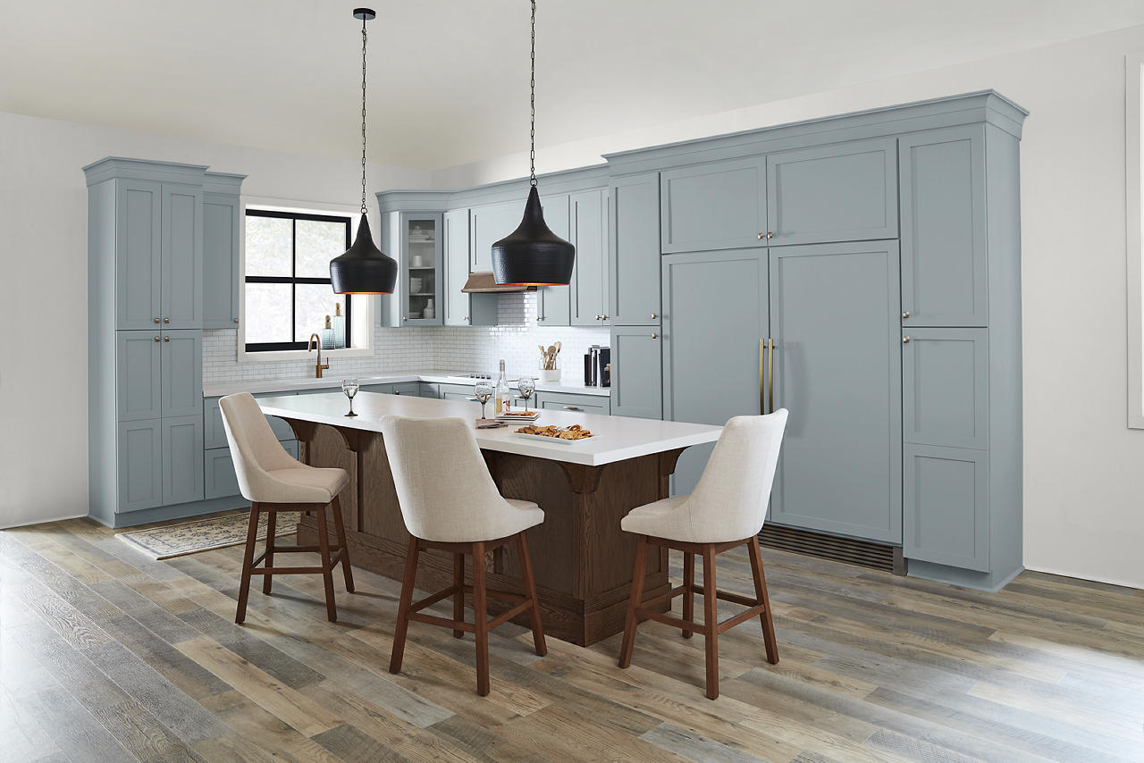 A kitchen with a popular soft blue paint colour on the cabinetry, brushed brass hardware, a dark wood island, warm wood flooring, black pendant lights, and cream bar stools.