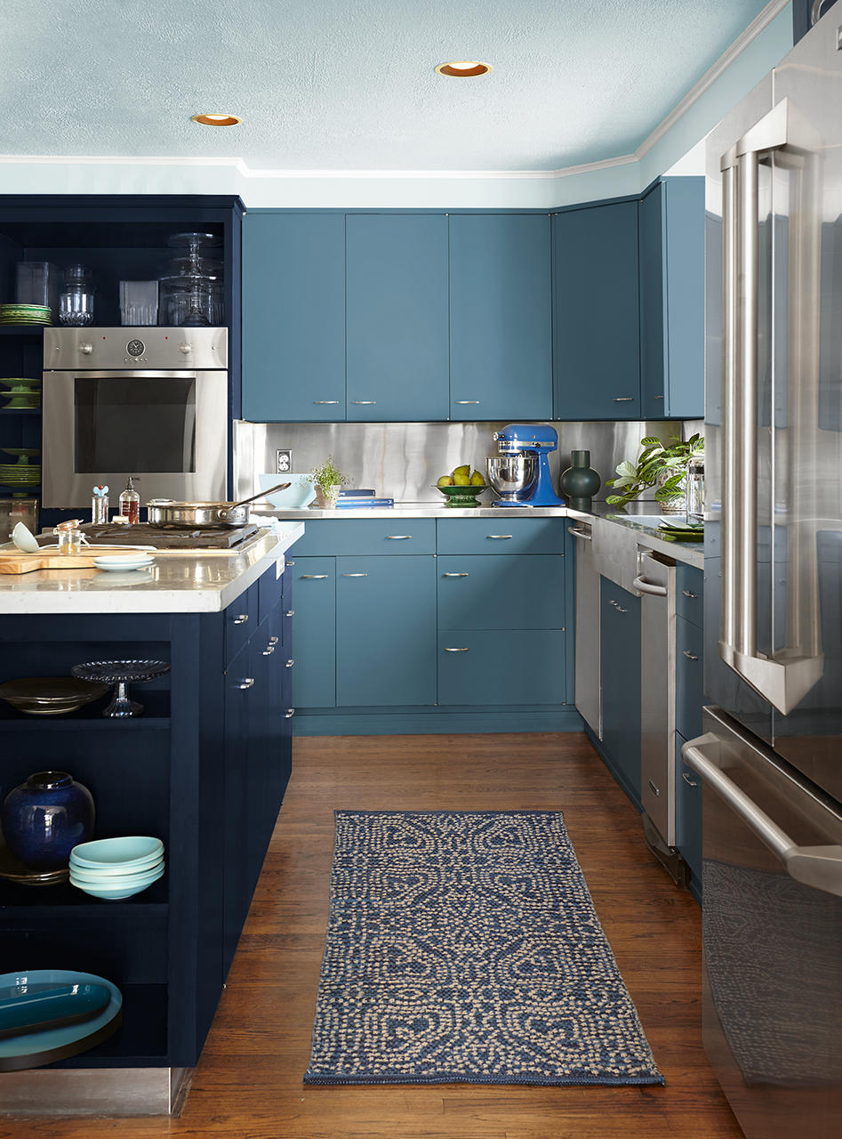 A blue colour-drenched kitchen with deep navy island cabinetry, softer blue wall cabinets, and a lighter coodinating blue ceiling that creates a bold, immersive look. A blue patterned runner and blue dishware continue the layered palette, while stainless steel appliances and warm wood floors add balance and contrast.