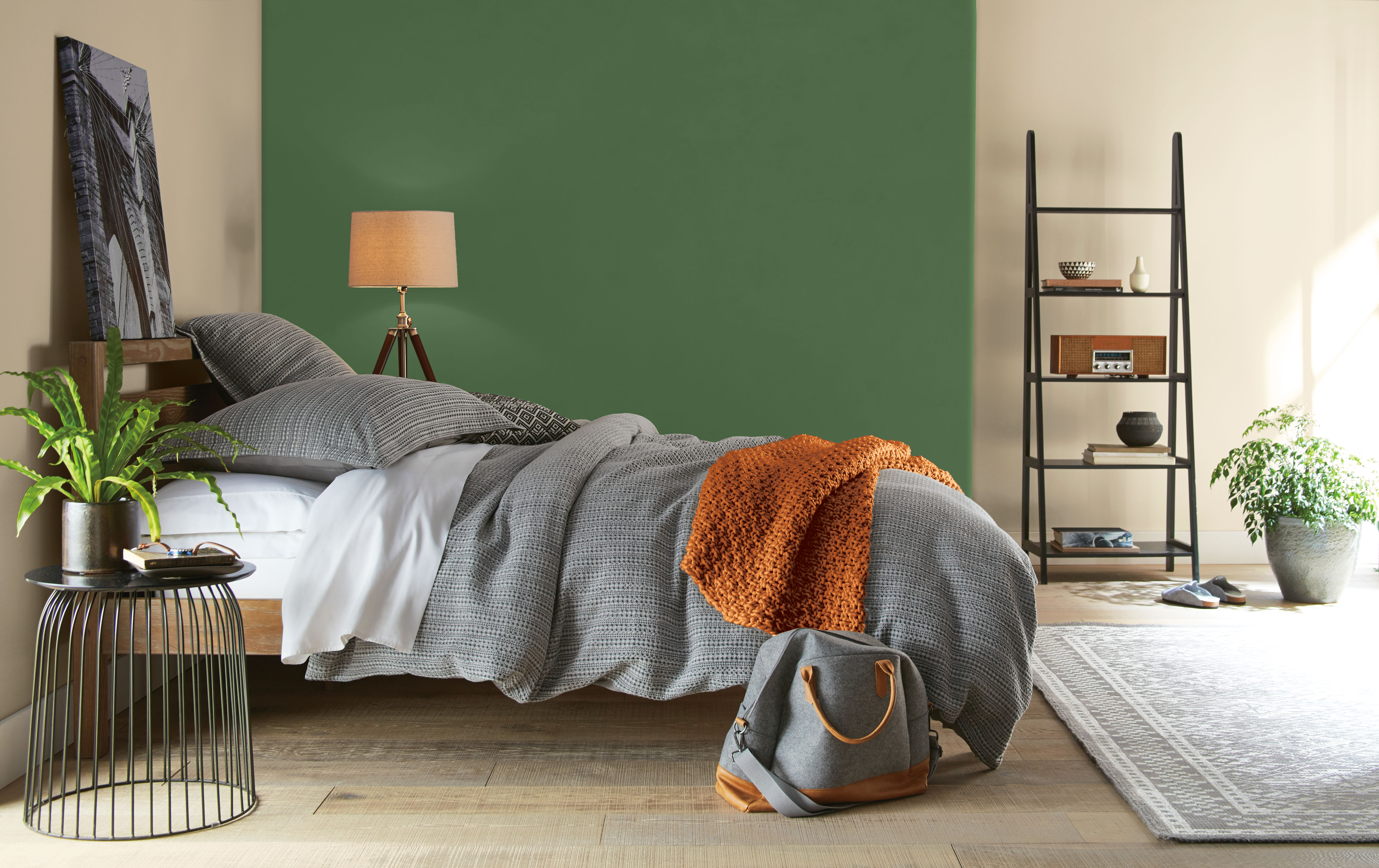 Bedroom with deep Garden Cucumber green accent wall, warm neutral walls, grey bedding, natural wood furniture, plants, and earthy orange accents.