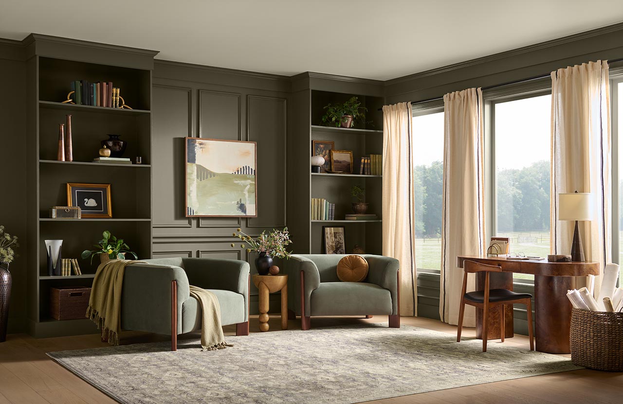 A cozy home office and study with walls painted in BEHR Grounded, featuring built-in bookshelves, two upholstered armchairs, a wooden desk, large windows with cream curtains, and warm natural light.