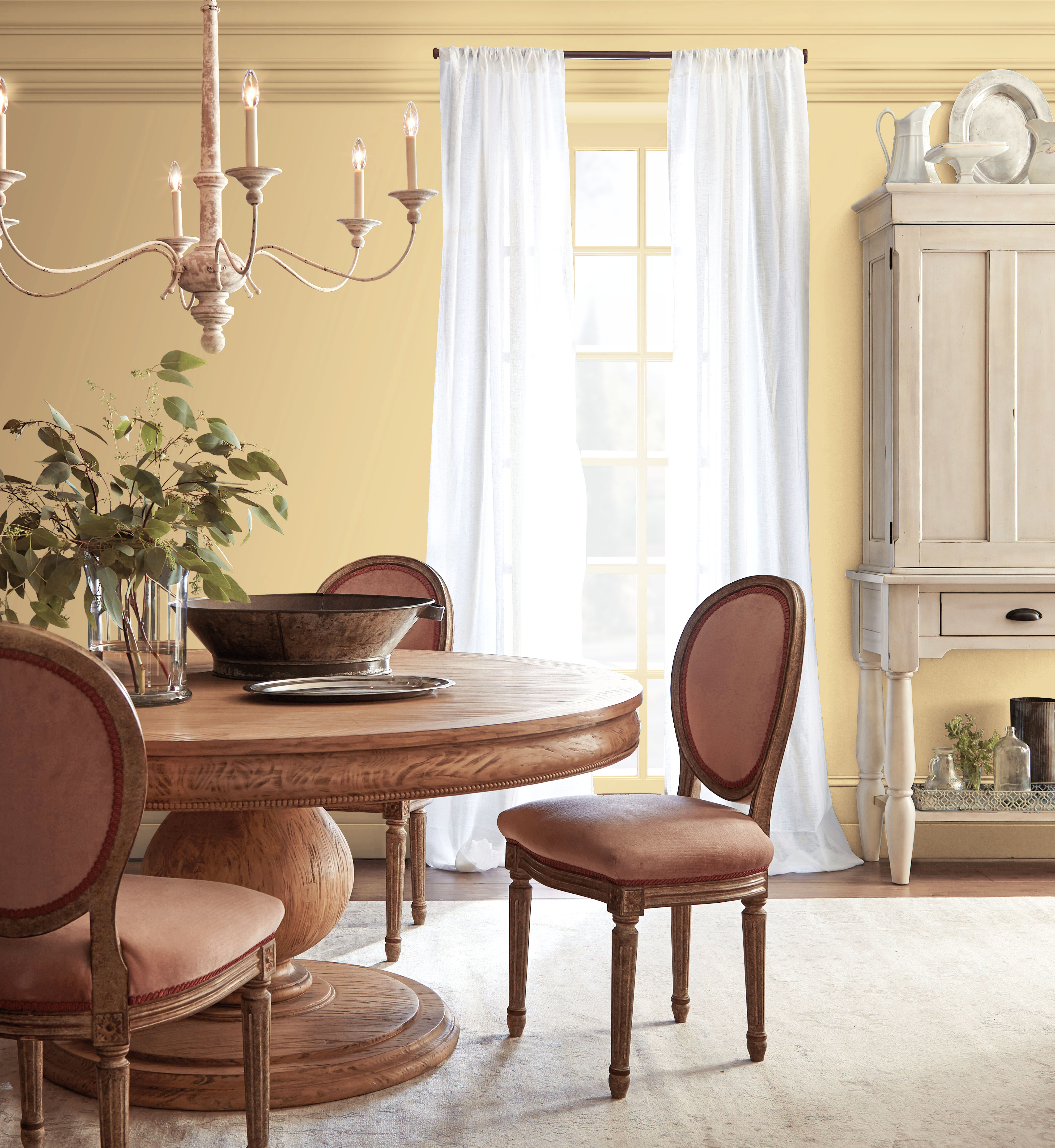 Dining room with Harmonious Gold wall paint, warm wood dining furniture, white curtains, traditional chandelier, and vintage-inspired décor for a romantic Regency-style dining room colour palette.
