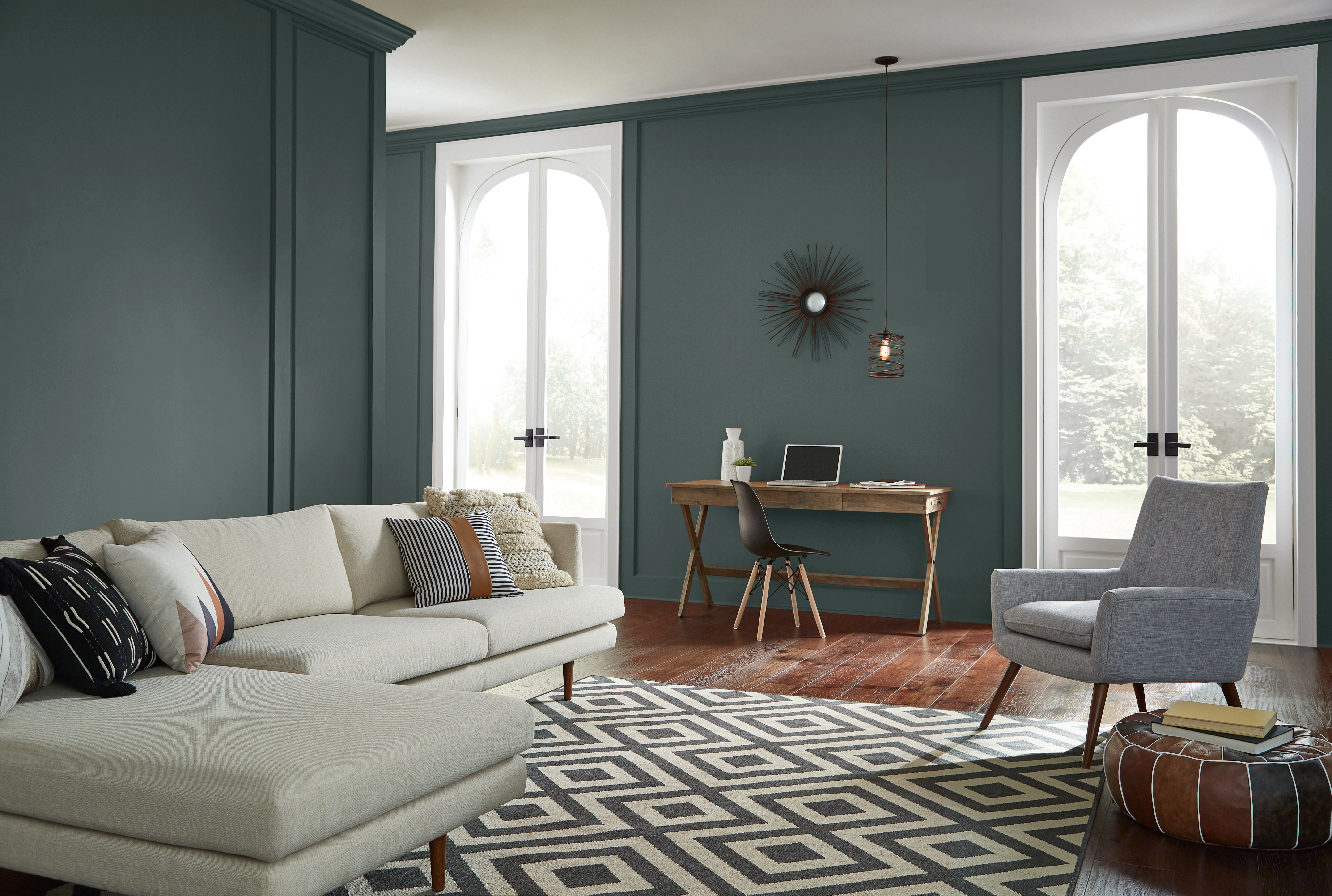 Open living room with dark green painted walls, small desk between tall windows, white sofa, patterned rug, grey chair, and wood flooring.