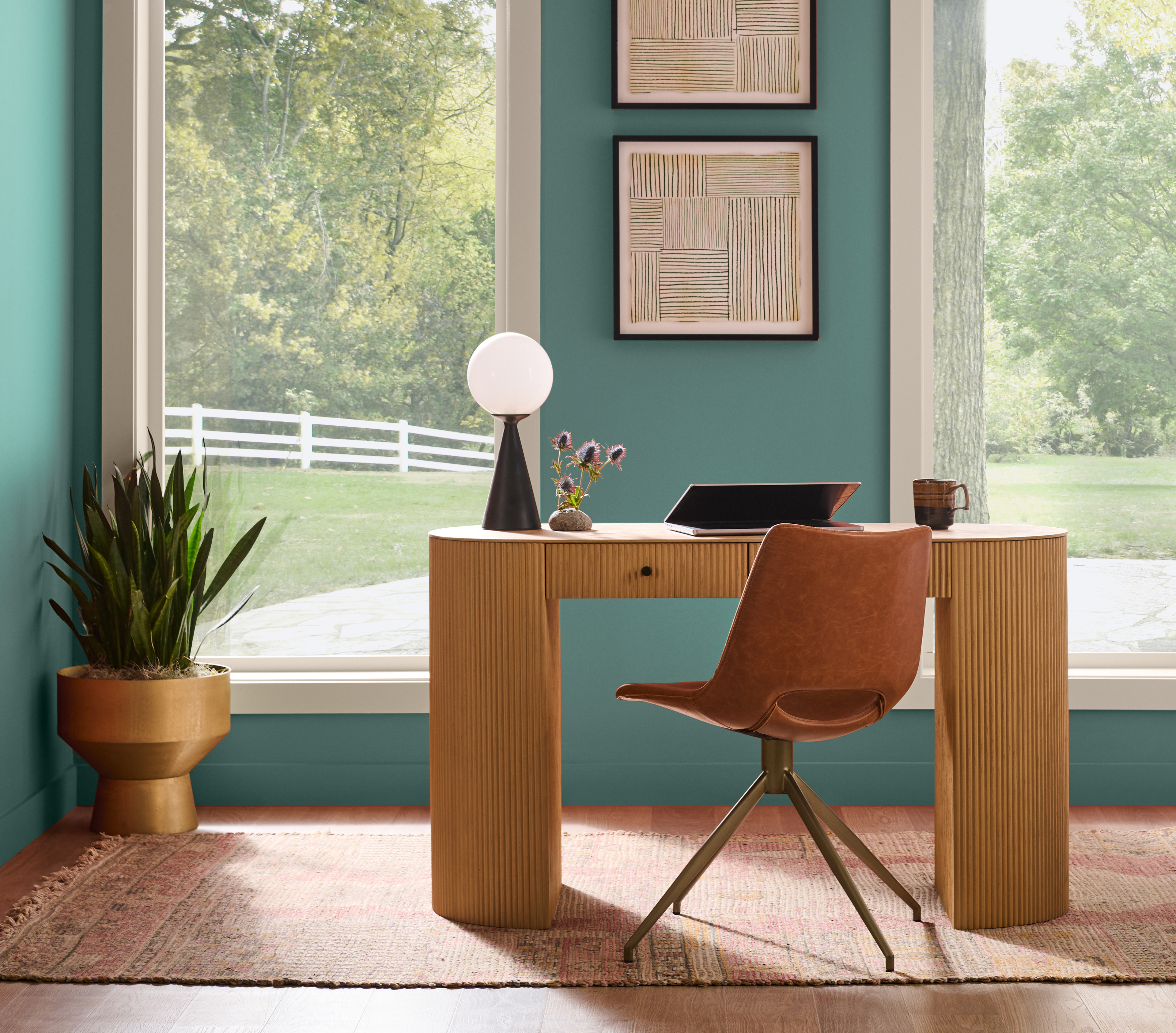 Home office nook painted in smokey jade hue with a small wood desk, windows, framed art, plants, and a chair in an open living area.