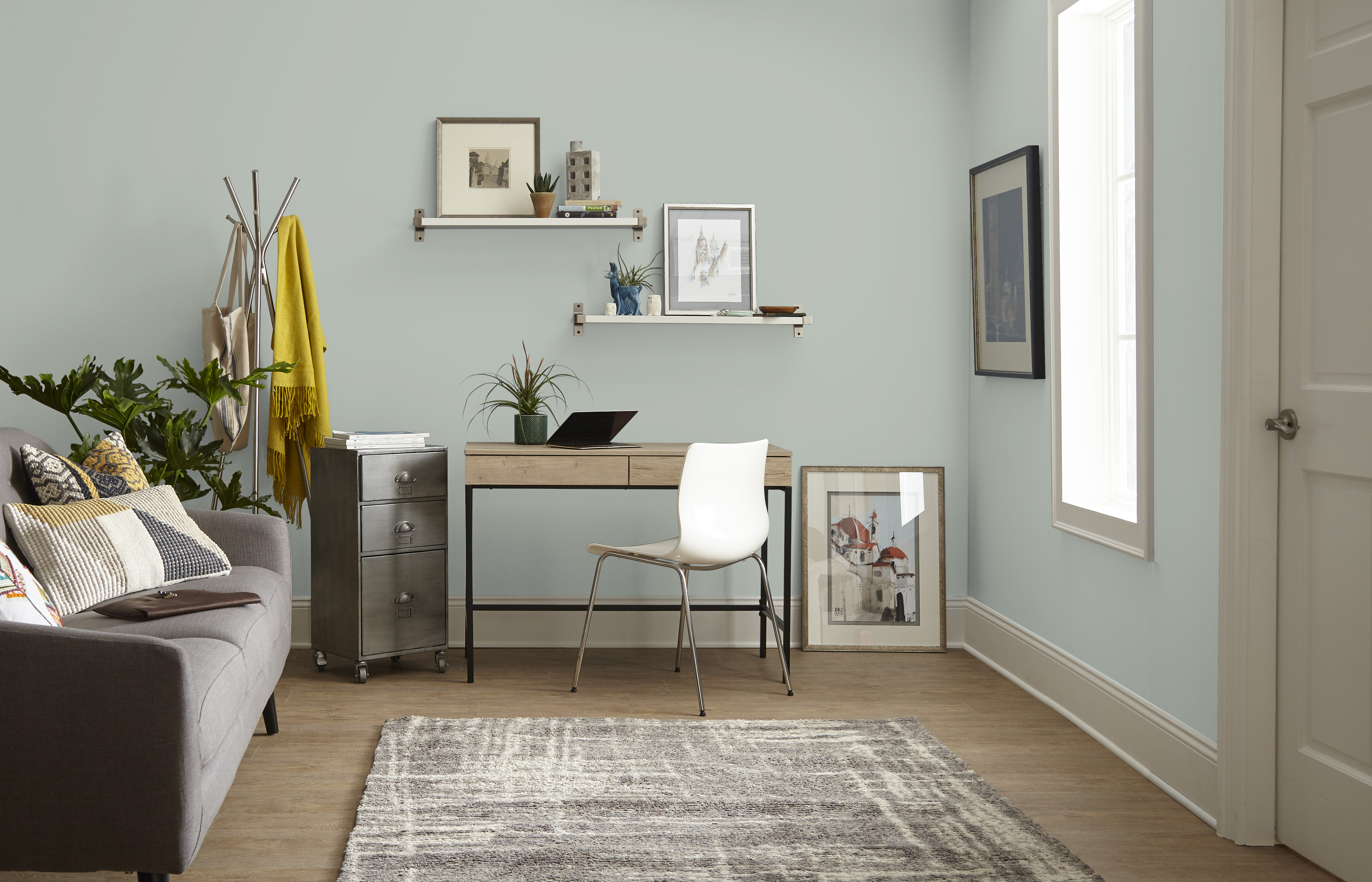 Small living room office with grey-green painted walls, compact desk, white chair, wall shelves, sofa, plants, and light wood flooring.