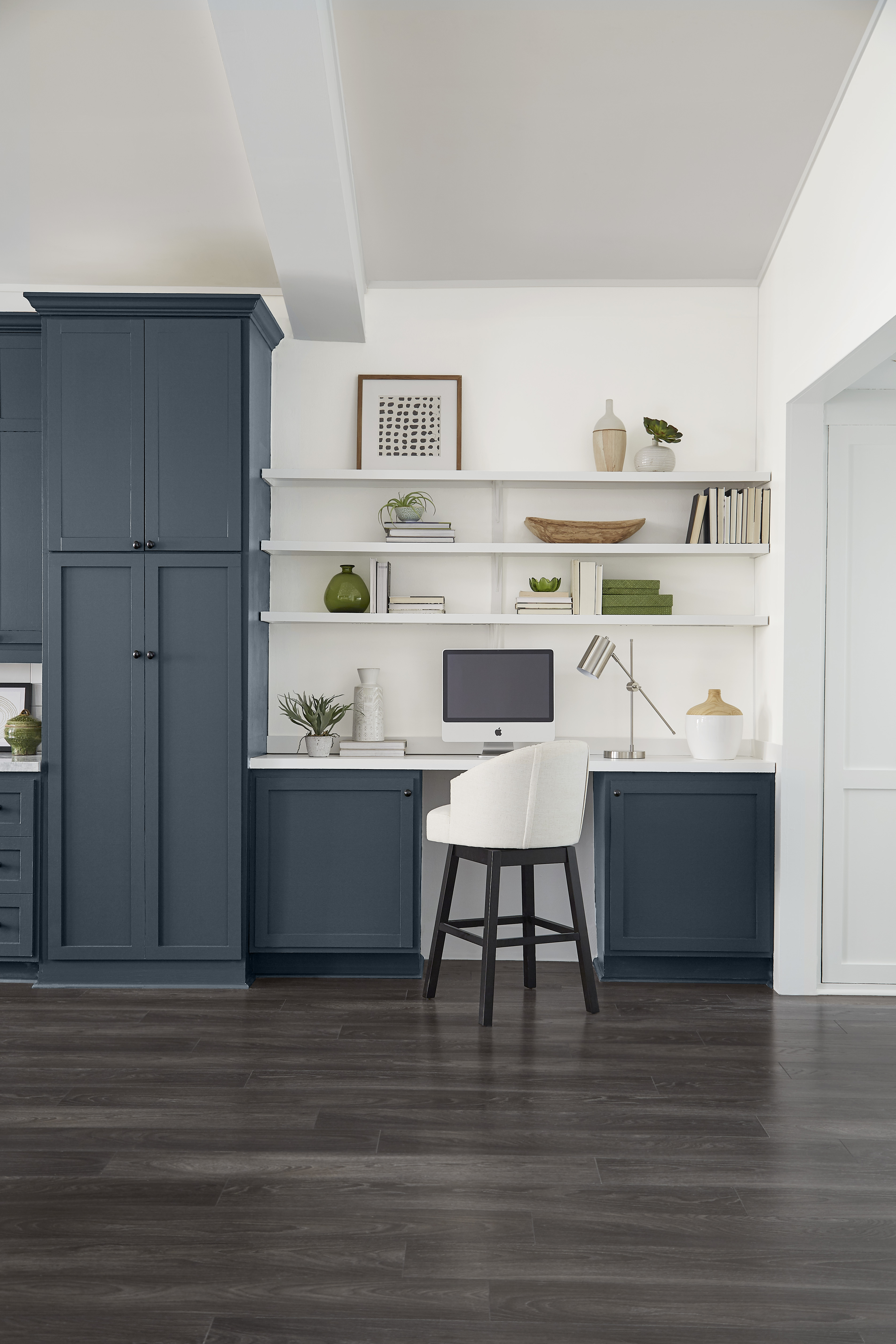 Kitchen-adjacent office area with deep blue-grey cabinetry, built-in desk, open white shelves, stool, computer monitor, and dark wood flooring.