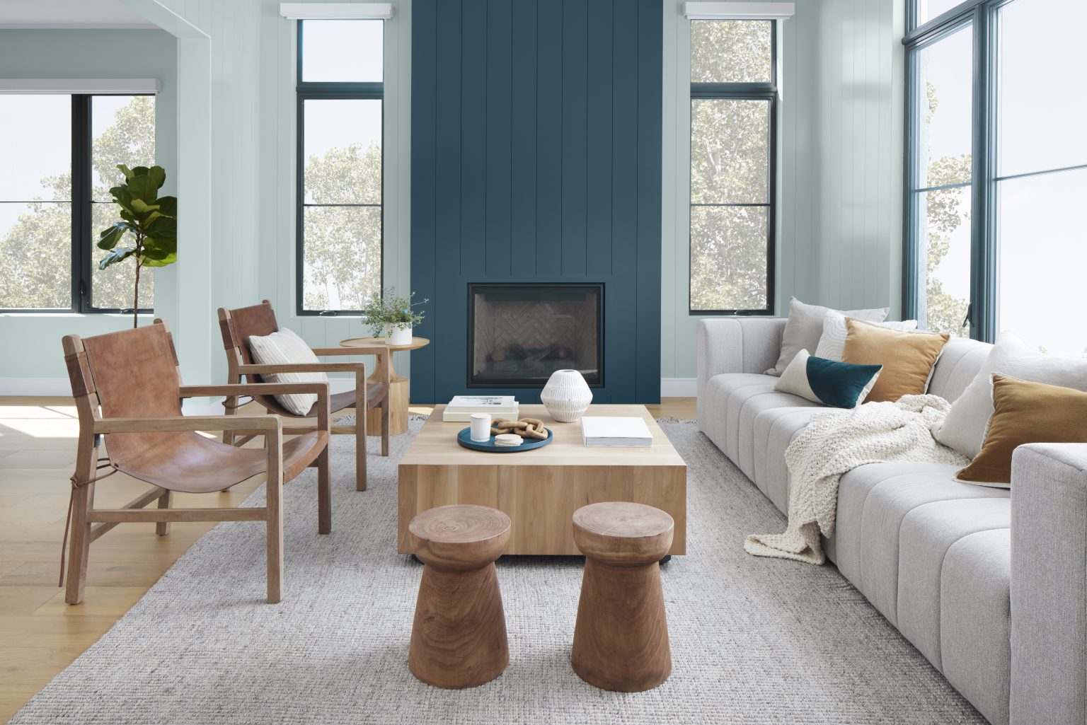 A bright, contemporary living room with walls painted Watery, a soft blue-green, and a vertical-paneled fireplace accent painted Nocturne Blue, a deep muted blue. Large black-framed windows, light neutral seating, natural wood furniture, leather sling chairs, and layered textures create an airy, relaxed space with a fresh coastal-modern feel.