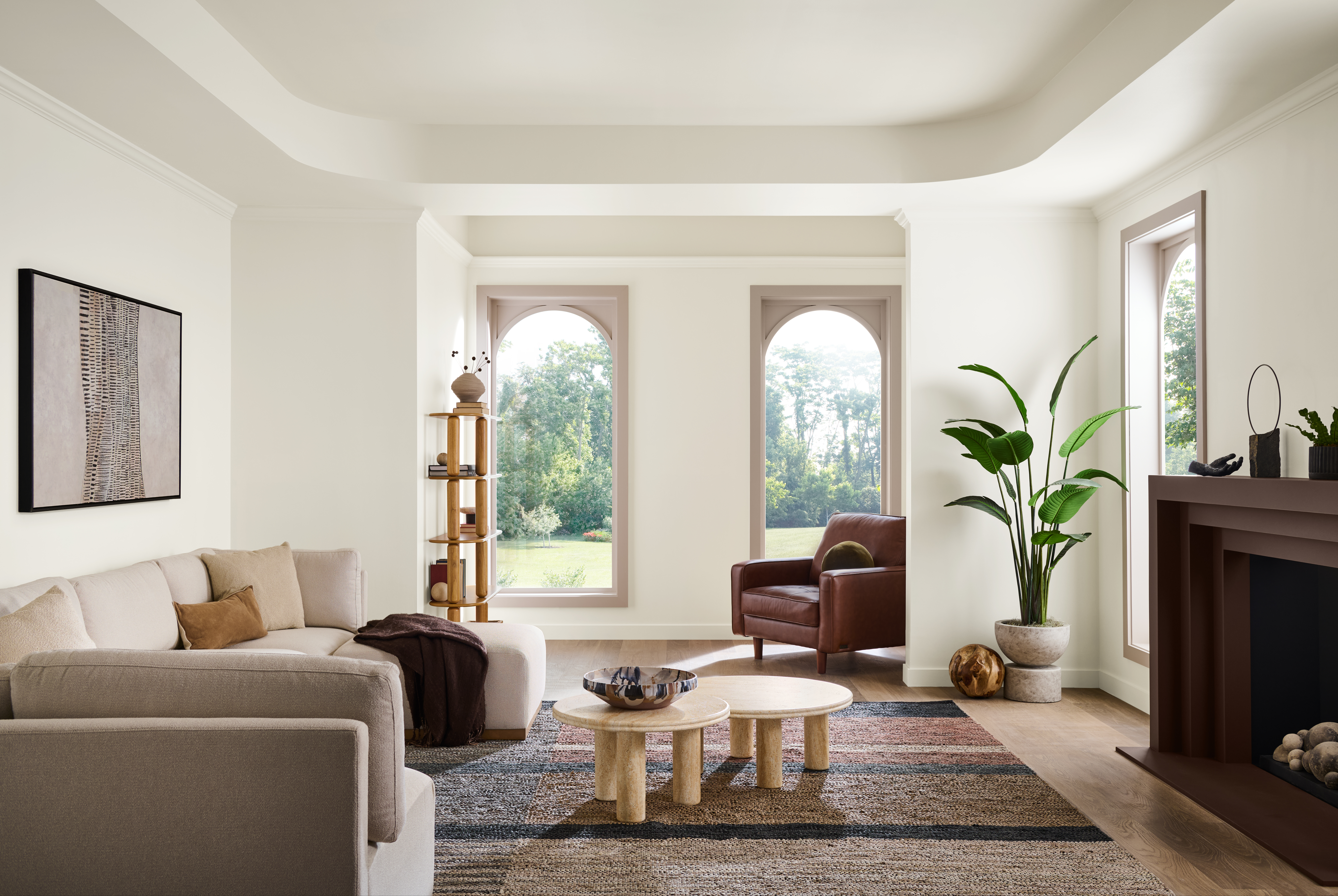 A warm, tonal living room with walls, ceiling, and baseboards painted Blank Canvas, window trim painted Mesa Taupe, and the fireplace mantel painted Baronial Brown. Layered warm neutrals, arched windows, soft upholstered seating, wood accents, and natural light create a relaxed, refined atmosphere.