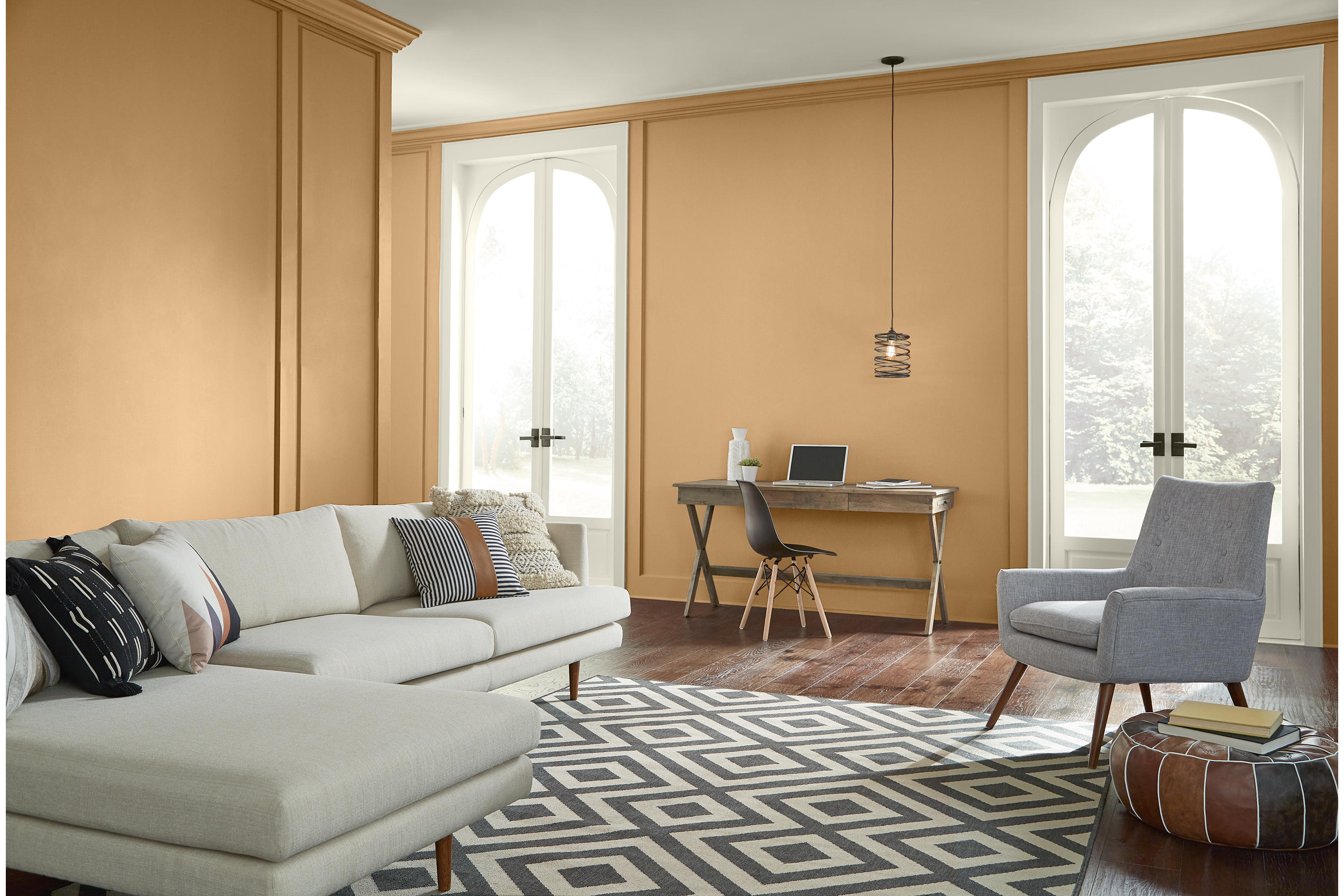 A bright living room with walls painted Cellini Gold and wood millwork painted Swiss Coffee, featuring tall arched windows, light neutral seating, a grey accent chair, wood flooring, and a geometric black-and-white rug.