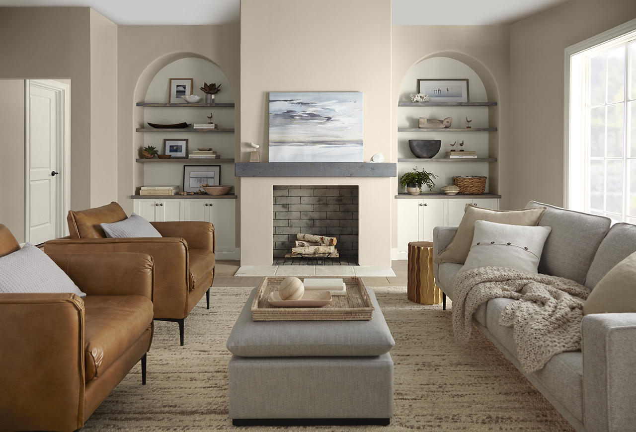 A warm, transitional living room with walls painted Even Better Beige and trim painted Blank Canvas. Camel leather armchairs, a soft grey sofa, arched built-in shelving, layered neutral textiles, and a simple fireplace create a comfortable, collected look. Abundant natural light and the warm beige-and-cream palette give the room an inviting, relaxed mood with subtle modern sophistication.