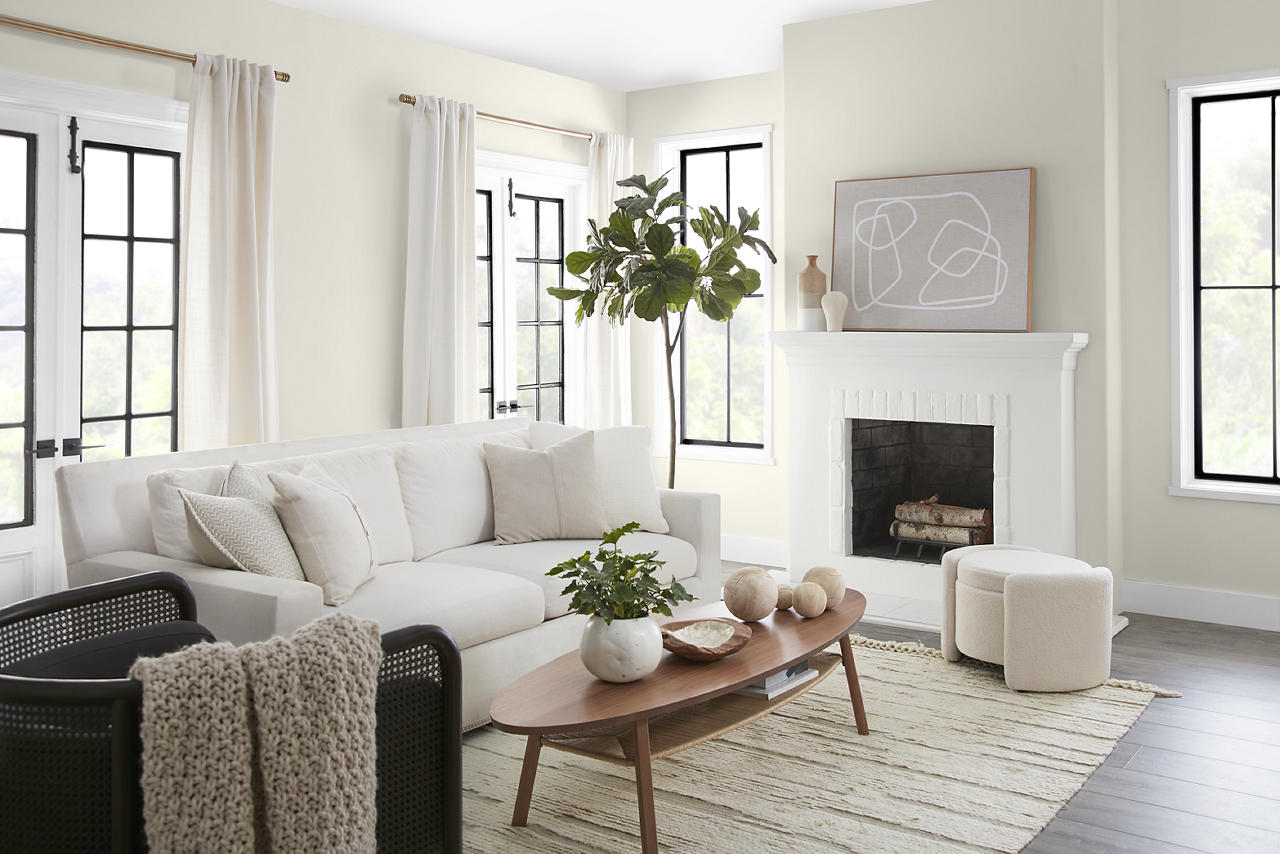 A bright, airy living room with walls painted Blank Canvas, a creamy soft white with a warm, welcoming undertone that feels balanced rather than yellow or stark. ULTRA PURE WHITE® highlights the trim and fireplace, while black-framed windows, white seating, light wood accents, and abundant natural light complete the space.