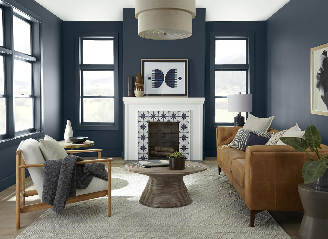 A casual living room with walls and trim painted Midnight Blue, a deep, moody navy blue, paired with ULTRA PURE WHITE® on the ceiling and fireplace. The space features a warm leather sofa, relaxed neutral seating, black-framed windows, natural wood accents, and a patterned tile fireplace surround.