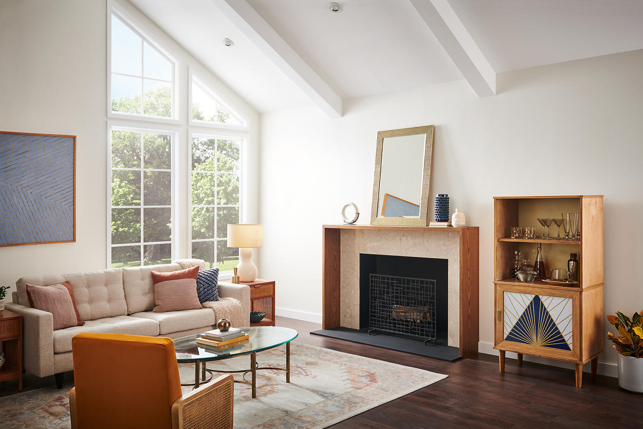 A bright mid-century modern living room with walls painted Diamonds Therapy and trim and vaulted ceiling beams painted ULTRA PURE WHITE®. Large windows fill the space with natural light, while warm wood furnishings, a streamlined fireplace, neutral seating, and a burnt orange accent chair add inviting mid-century modern flair.