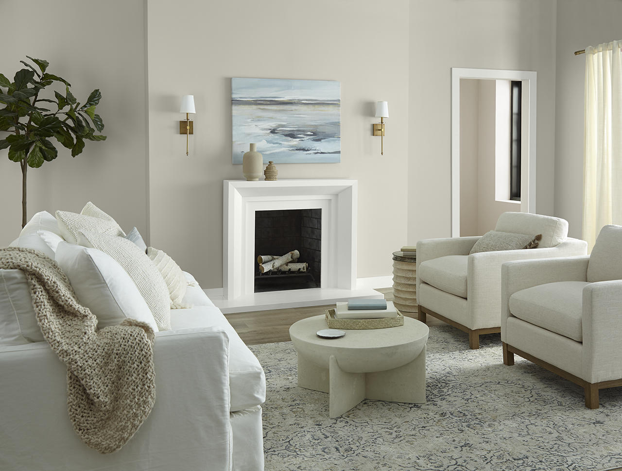 A bright, relaxed living room with a subtle coastal feel, featuring walls painted Tranquil Gray, a soft warm grey, a hallway wall painted Even Better Beige, and trim painted Whipped Cream. Exposed wood ceiling beams, white upholstered seating, a simple fireplace, natural textures, and soft daylight create an airy, comfortable atmosphere.