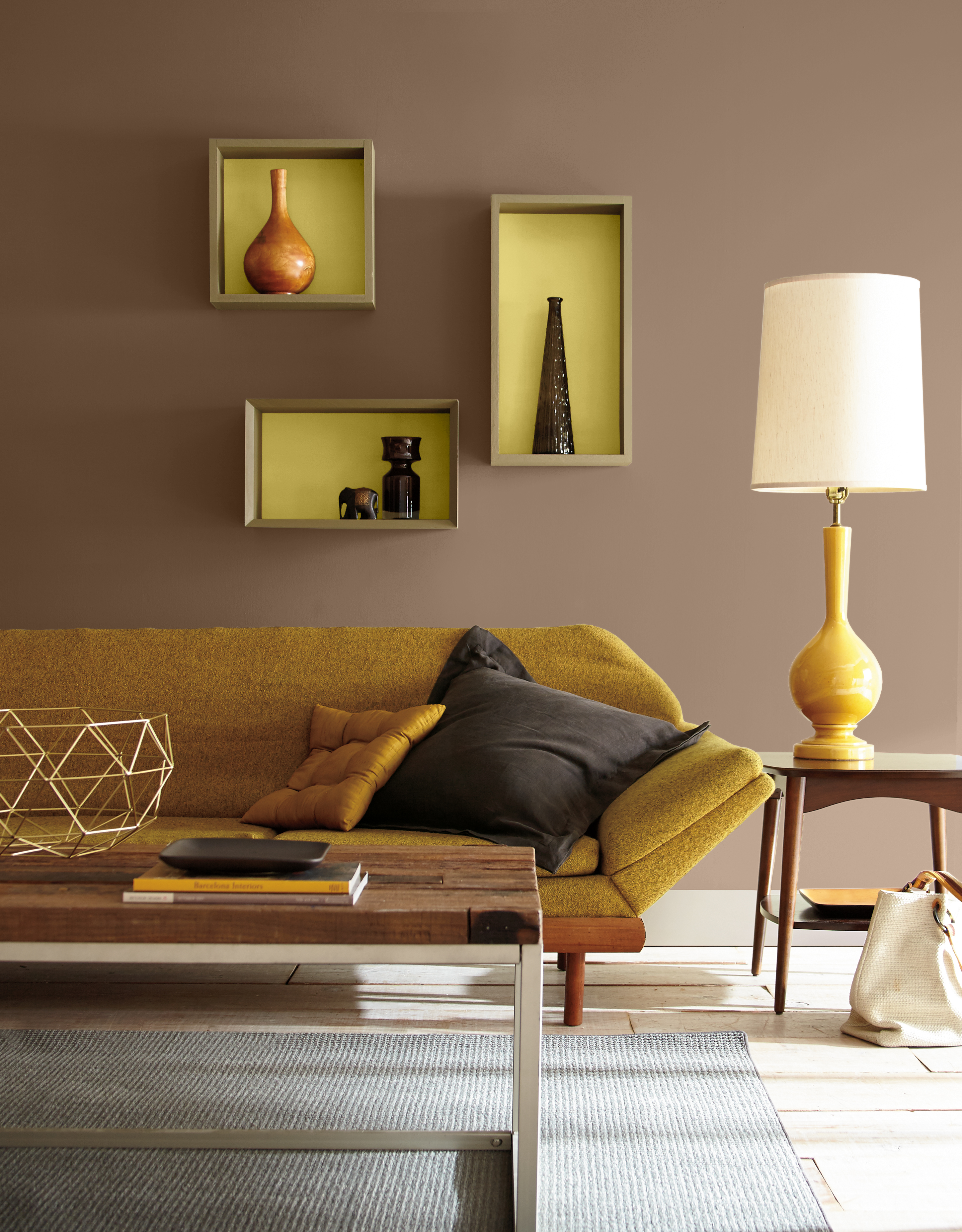 A retro-inspired living room with walls painted Joshua Tree, a warm earthy brown, accented by Starfruit, a lively yellow-green, inside the recessed display niches. A low-profile mustard sofa, vintage-style table lamp, sculptural wood and metal coffee table, geometric décor, and mid-century furnishings create a nostalgic, collected look.