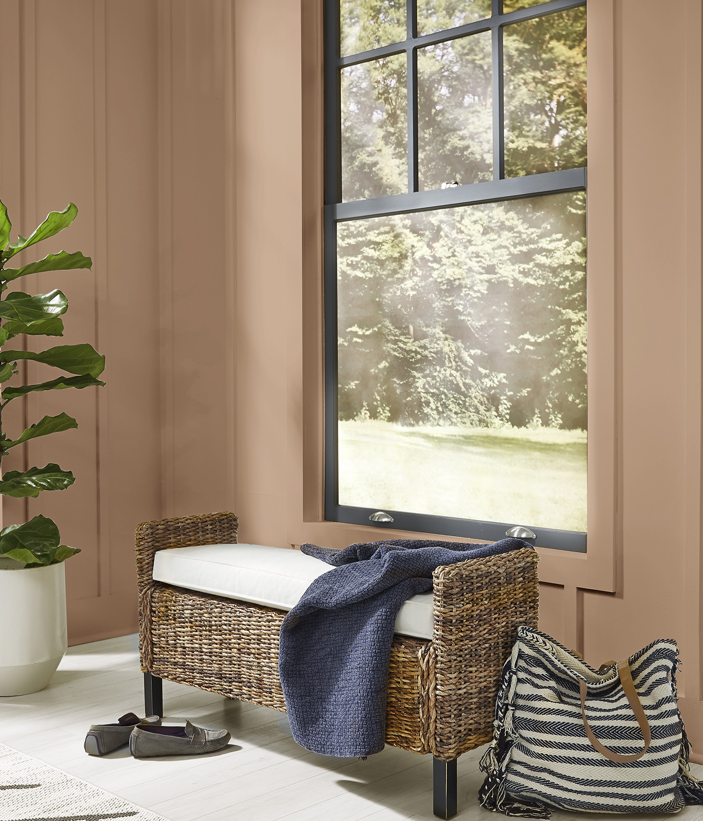 A wicker bench with a white cushion and blue blanket sitting in front of a tall window with walls painted in a beige paint color. The décor is casual and comfortable.